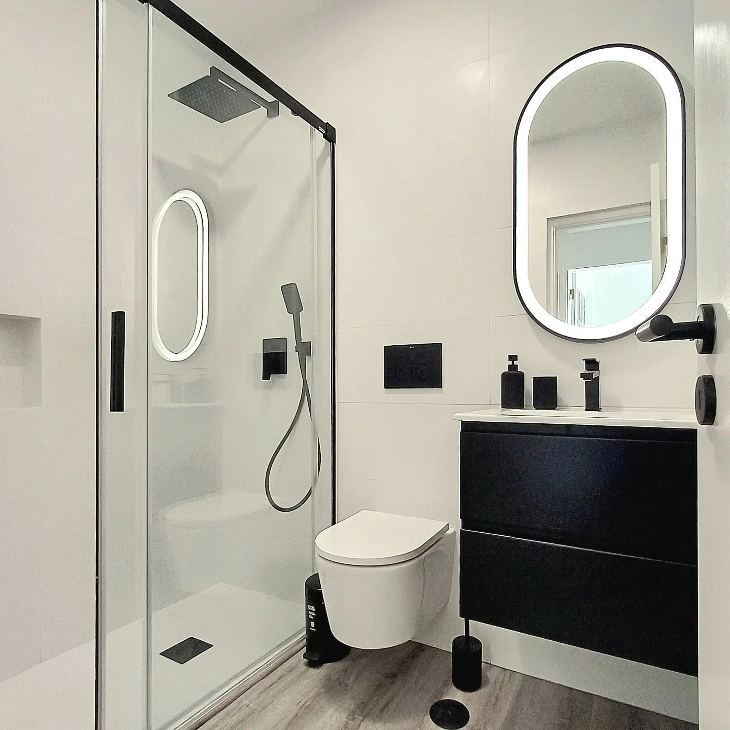 Banheiro moderno com box de vidro, vaso sanitário, espelho oval com iluminação, pia preta com utensílios preto, parede branca e piso de madeira clara.