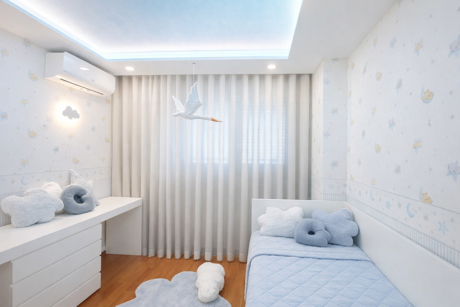 Decoração de Quarto de bebé em Lisboa, decorado em tons claros, predominando o azul bebé, com papel de parede figurativo, nuvens e estrelas na parede, almofadas em formato de nuvem, cortina de tecido branco e um mobile de cegonha sobre a cama.