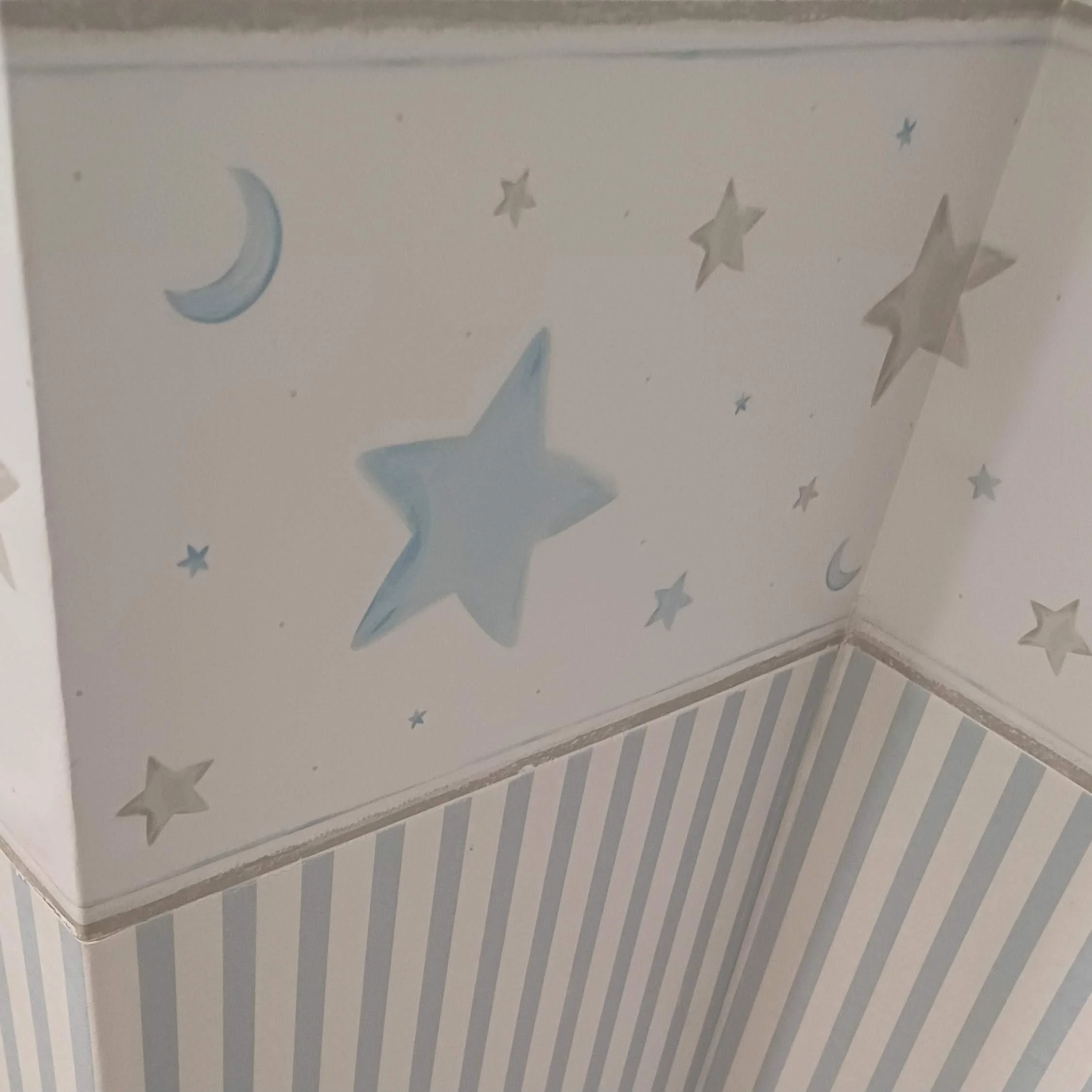 Parede de quarto infantil decorada com estrelas e luas em tons de azul, branco e cinza, com teto de listras azul e branco