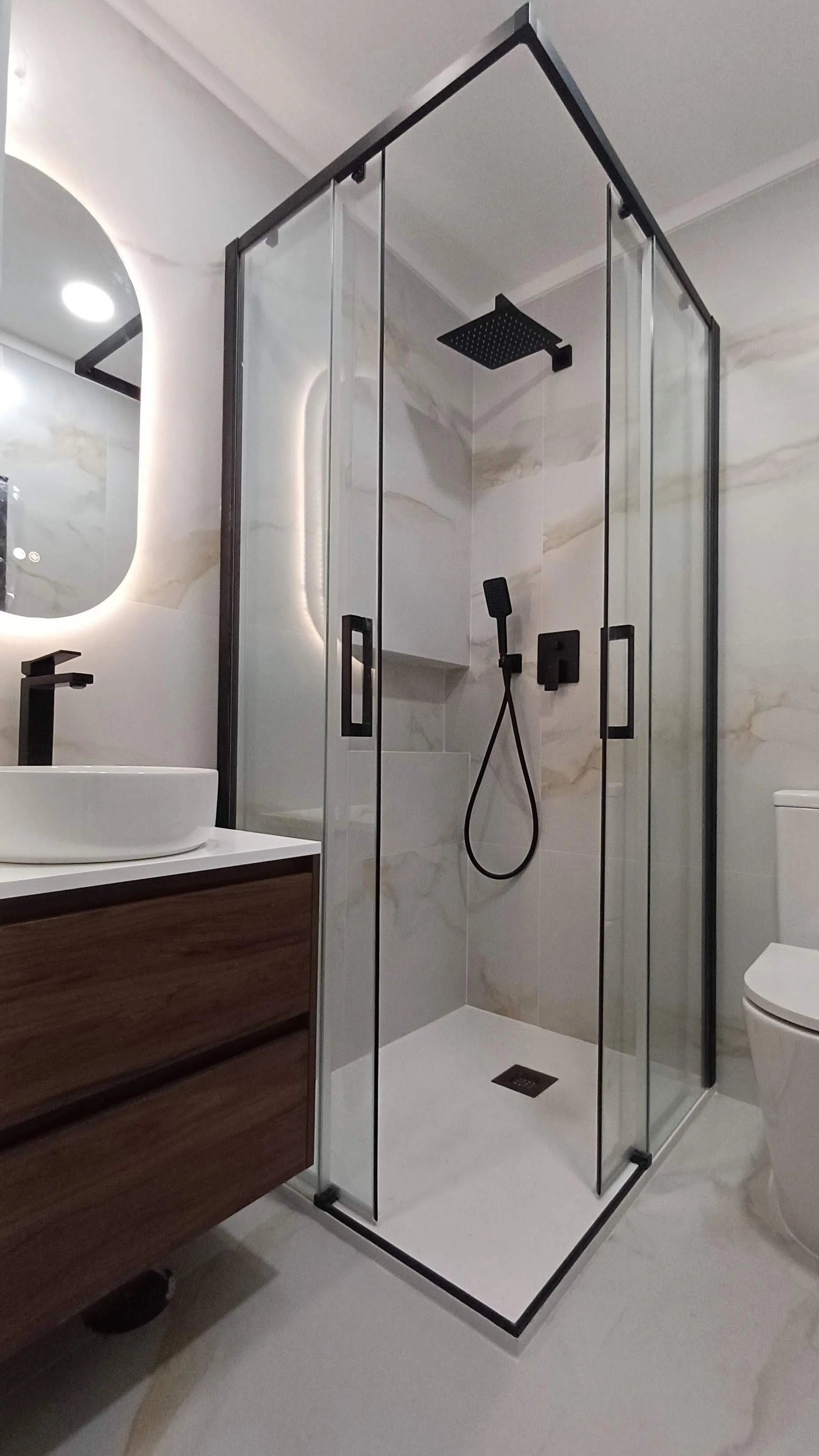 Banheiro com chuveiro de vidro preto, pia de Eva com torneira preta, espelho oval com iluminação embutida e vaso sanitário branco, decorado com azulejos brancos com veios dourados.