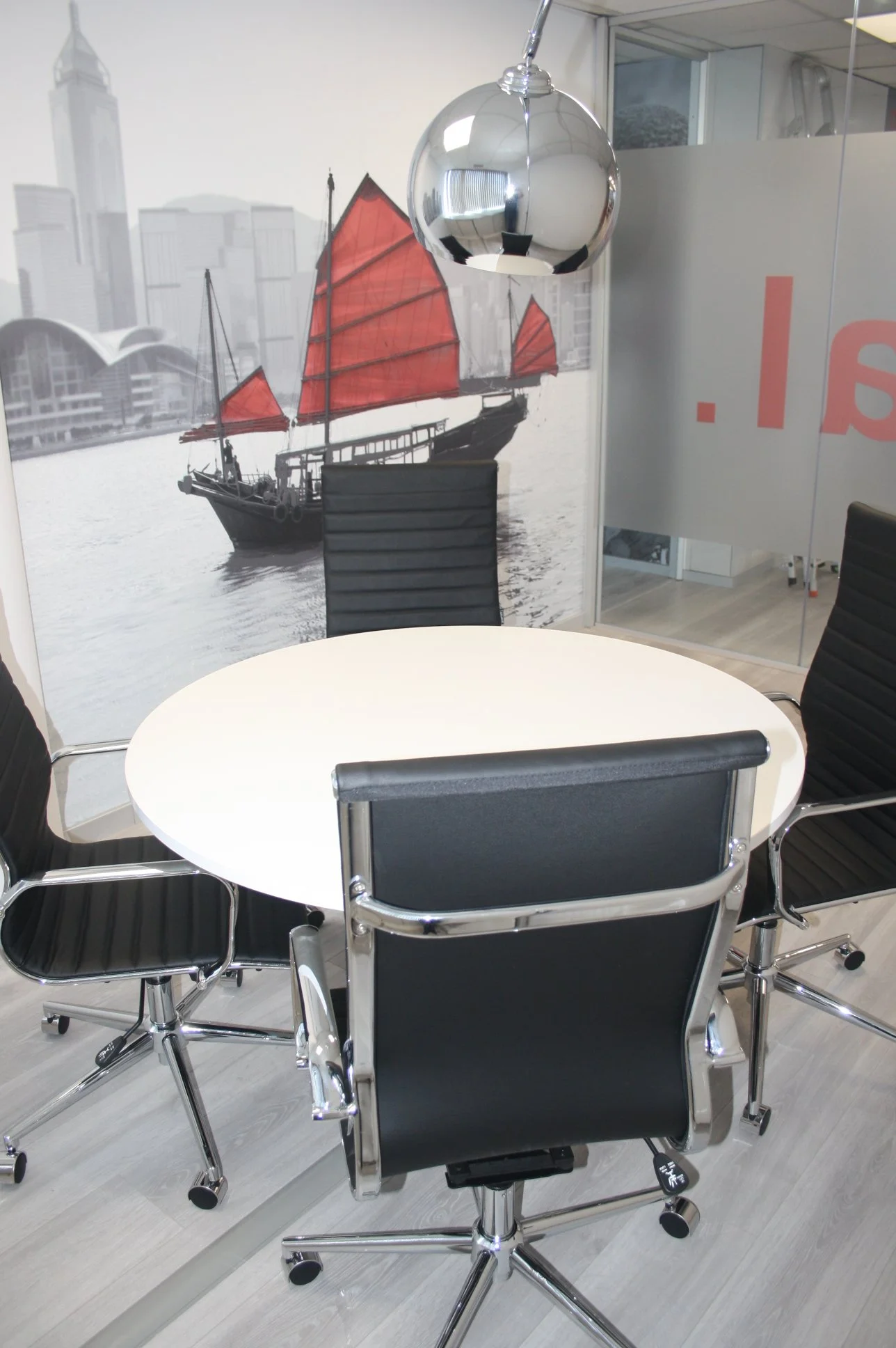 Reunião com mesa branca, cadeiras de escritório pretas com detalhes de metal, parede com pôster de um barco com velas vermelhas, luminária espelhada pendurada no teto, parede de vidro com reflexo de escritório.