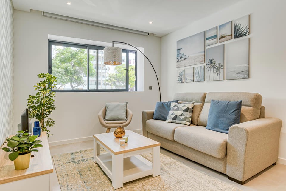 Sala de estar com sofá bege, dois travesseiros azuis e um decorativo com padrão geométrico, uma poltrona giratória, uma mesa de centro branca com objetos decorativos, uma parede com quadros de praias e natureza, uma janela grande com vista para árvor