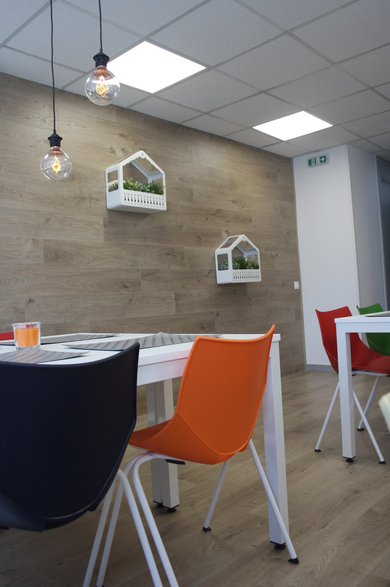 Sala de jantar com mesa branca, cadeiras coloridas (preta, laranja e vermelha), parede de madeira decorativa com prateleiras brancas em formato de casas com plantas, luminárias de teto com lâmpadas expostas, piso de madeira, porta de saída de emergên