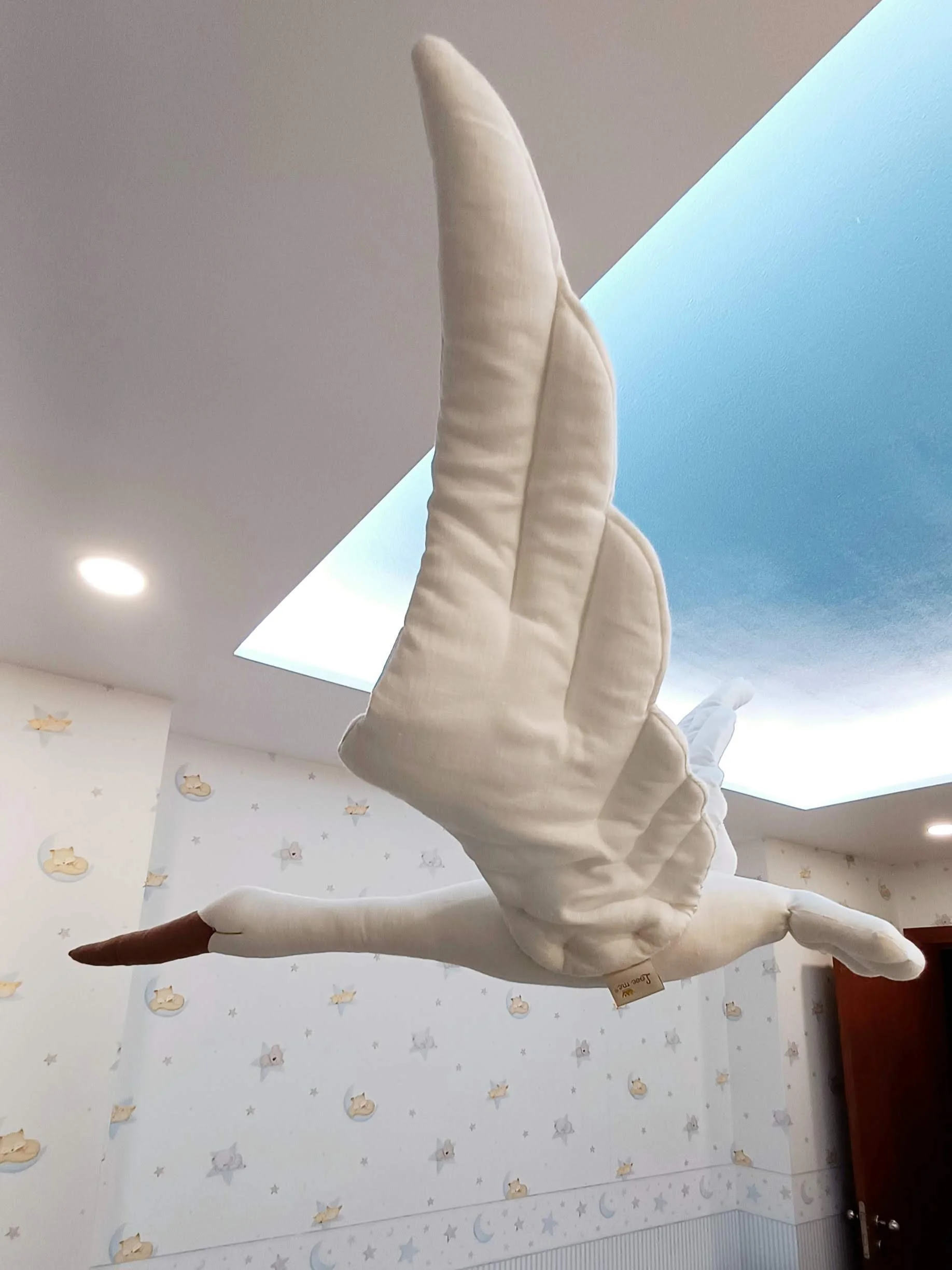 Brinquedo de pelúcia de um cisne com corpo branco e bico marrom, pendurado no teto de um quarto decorado com papel de parede de desenhos de nuvens e estrelas.