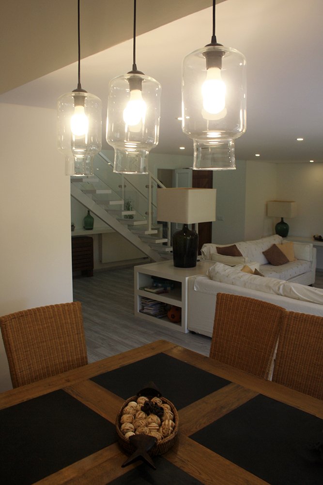 Sala de estar com teto iluminado, sofá branco com almofadas, mesas laterais, lâmpadas de vidro penduradas, mesa de jantar de madeira com travessas de diferentes cores e uma escada ao fundo.