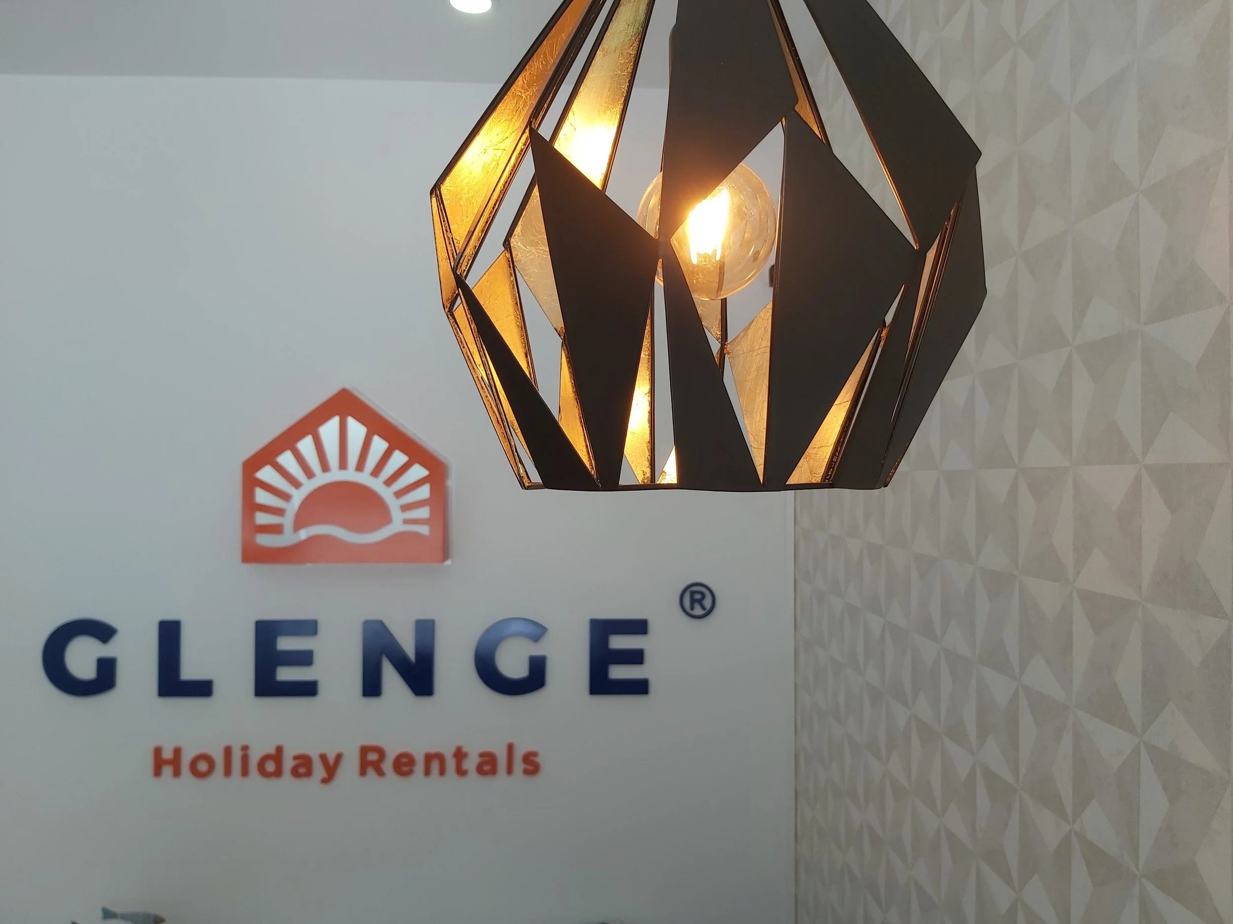 Projeto de Design de Interiores/Remodelação.
Pormenor de área de recepção da Glenge Holiday Rentals. Em primeiro plano um candeeiro pendente sobre o balcão. Ao fundo o logo Glenge em parede frontal.