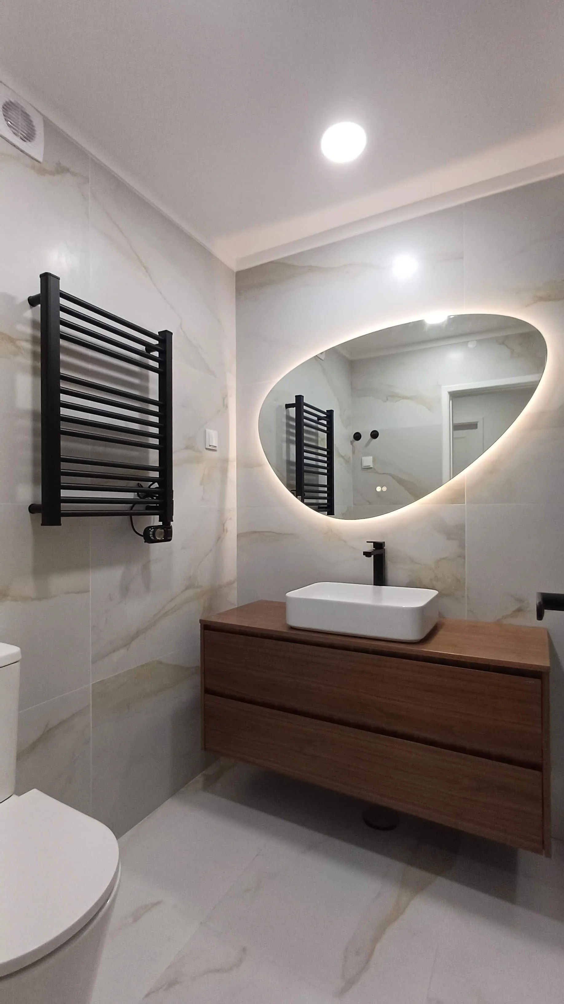 Banheiro moderno com parede de mármore, espelho oval com iluminação, pia de mesa de madeira e torneira preta, toalheiro preto e vaso sanitário branco.