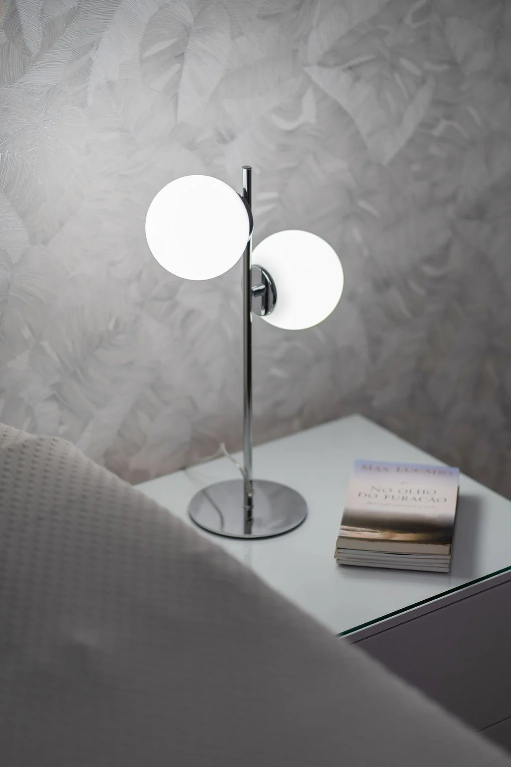 Luminária moderna com duas lâmpadas brancas em uma mesa ao lado de um livro.