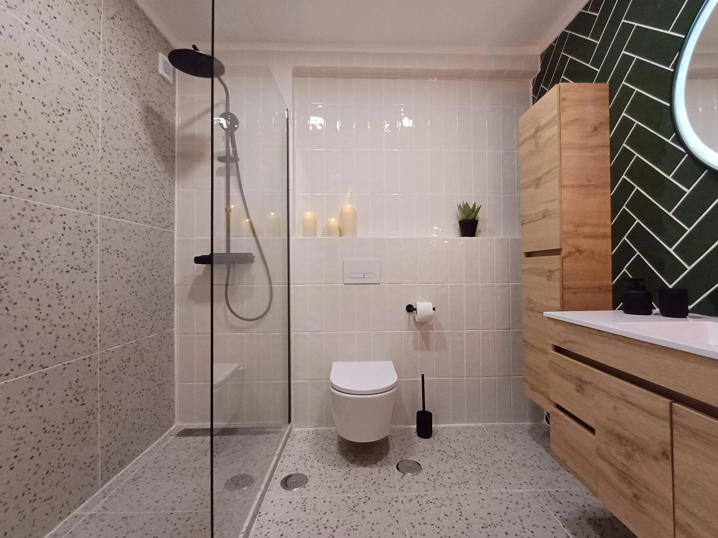 Banheiro moderno com parede de azulejos brancos, área de banho com vidro e chuveiro preto, vaso sanitário branco, vaso de plantas, banheiro com piso e paredes de azulejos com textura abrasiva e escura, iluminação suave com velas, armário de madeira c