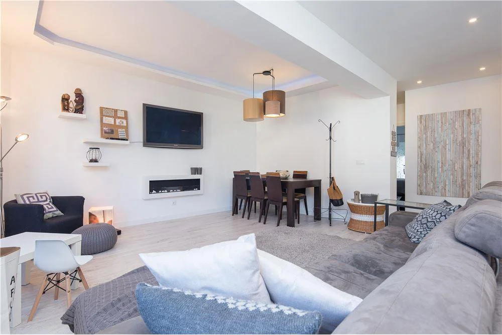 Sala de estar moderna com sofá cinza, televisão na parede, mesa de jantar com seis cadeiras, iluminação suave e decoração minimalista.