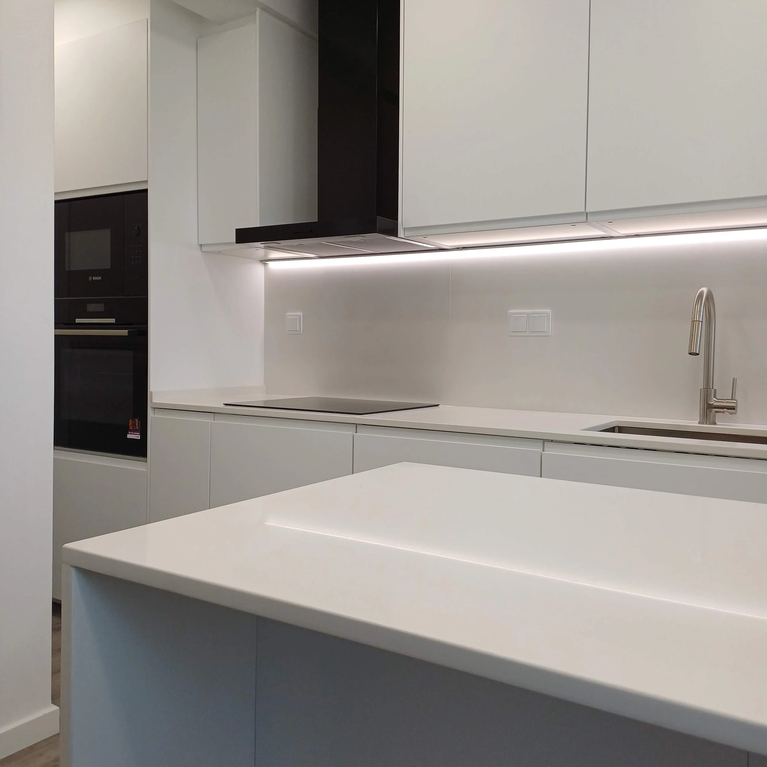 Remodelação de Cozinha no Parque das Nações, Lisboa. Cozinha com Decoração Preto/Branco, Pavimento vinílico imitação de madeira cinza, armários brancos mate com puxador integrado, tampo e back-splash em silstone branco. Equipamentos em cor preta.