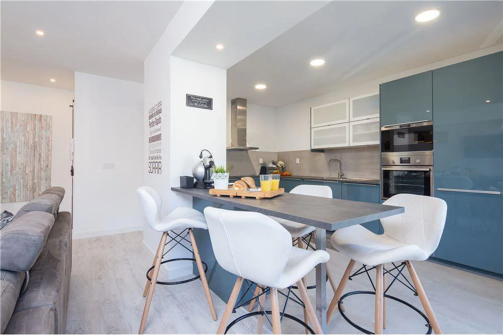 Cozinha moderna com armários azuis, bancada de granito, mesa de jantar com quatro cadeiras brancas, eletrodomésticos embutidos e iluminação de LED no teto.