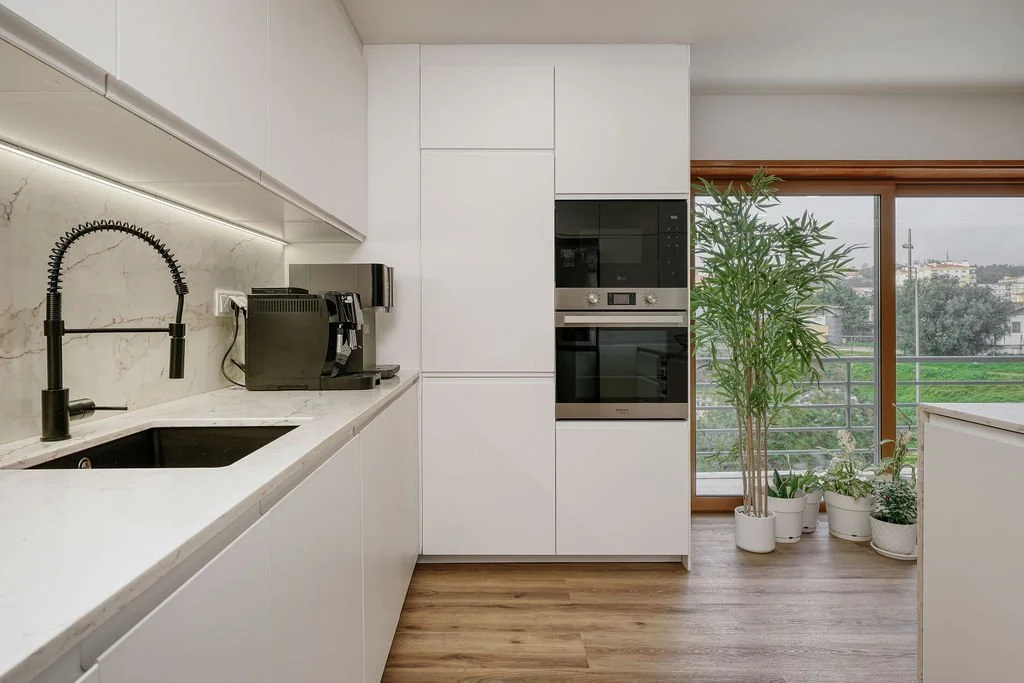 Cozinha moderna com guarda-roupa branco, pia preta, máquina de café, forno embutido, janela com vista para um espaço ao ar livre com árvores, e várias plantas em vasos brancos.