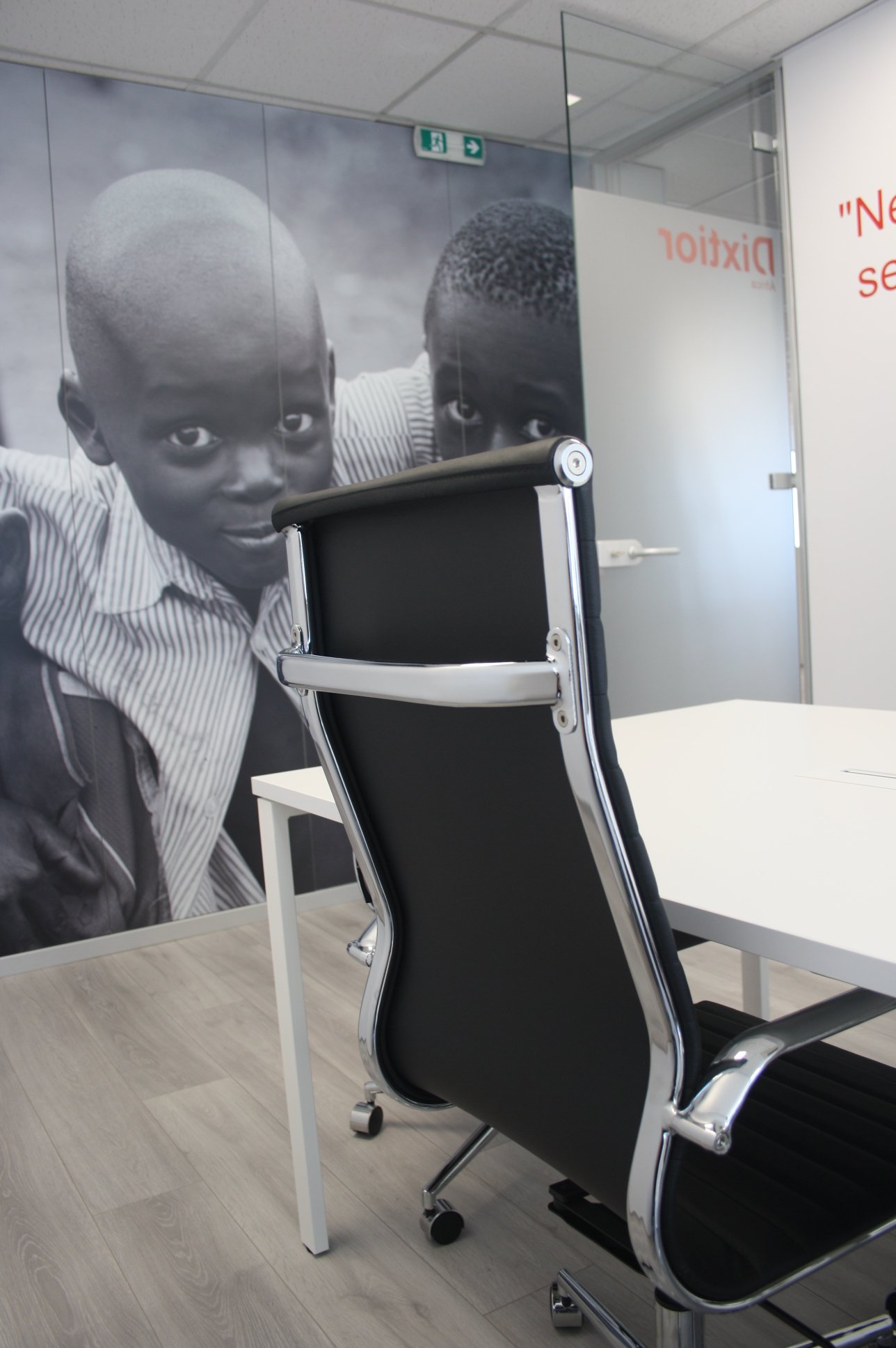 Sala de reunião com uma cadeira de escritório preta e branca, parede com grande painel fotográfico preto e branco de duas crianças jovens, fundo de parede branco com texto vermelho e uma porta de vidro com uma placa de evacuação.