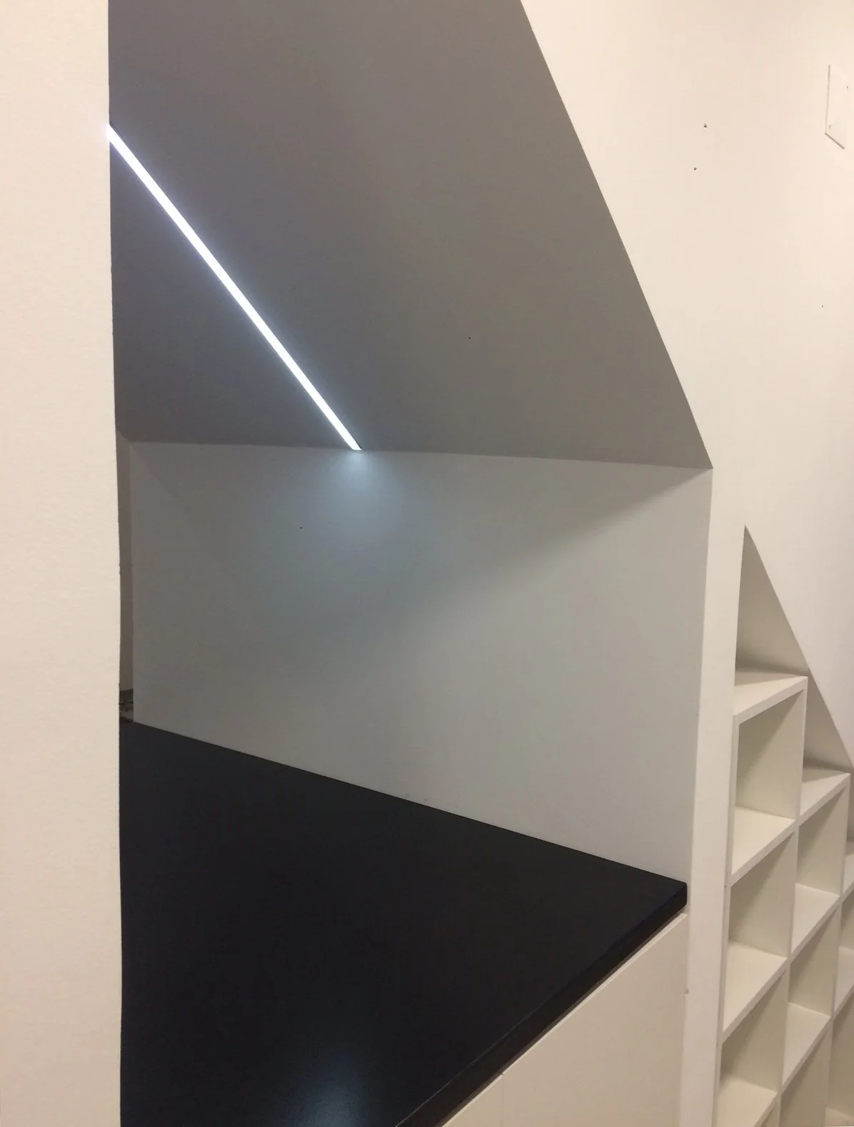 Imagem de um espaço interior com teto inclinado, iluminação de luz branca e uma superfície preta, com uma parede branca com nichos em formato de cubo.