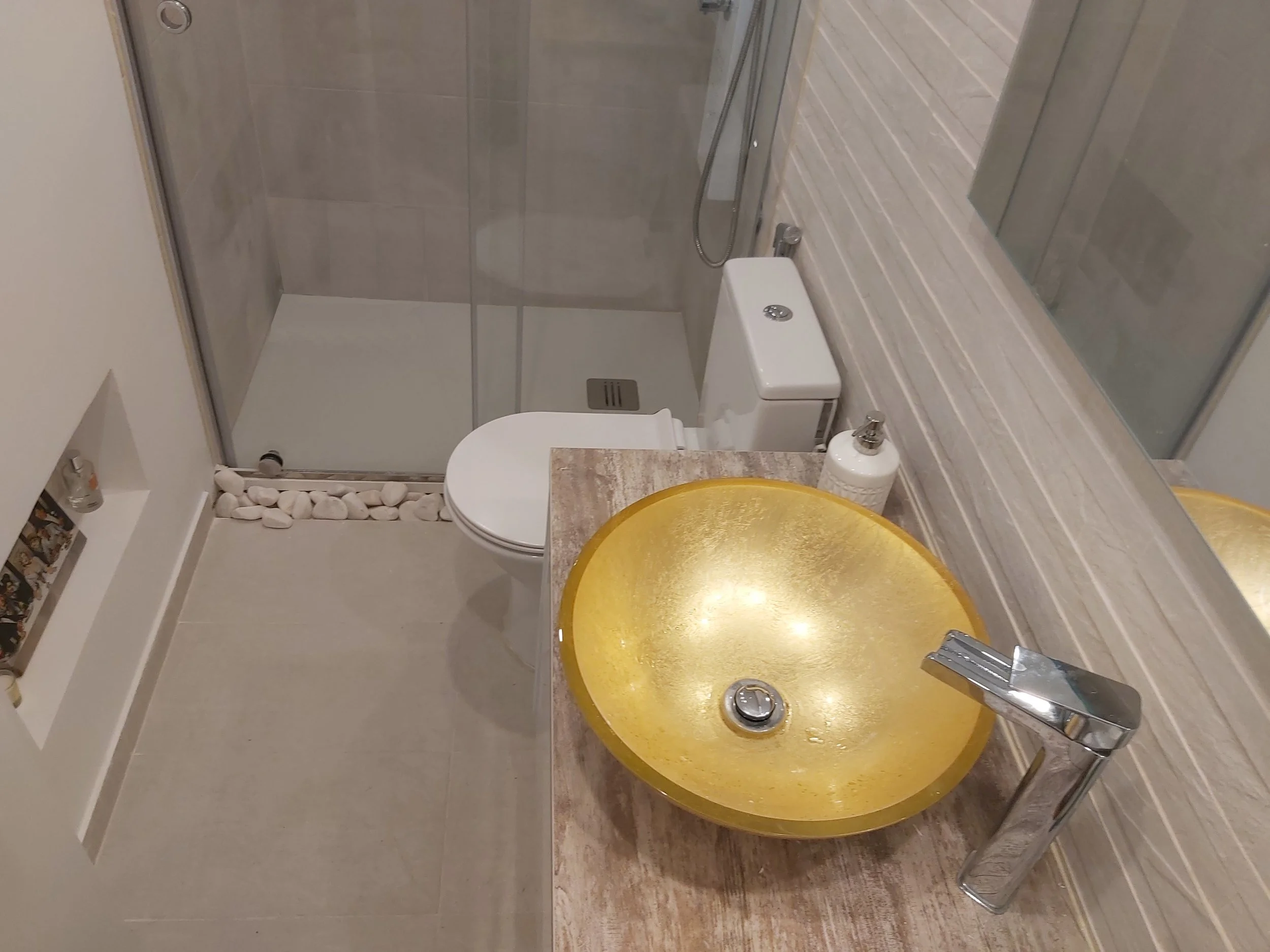 Banheiro com pia de vaso em vidro dourado, vaso sanitário, box de banheiro de vidro e parede de revestimento em tom bege com detalhes de pedras de rio na base da banheira.