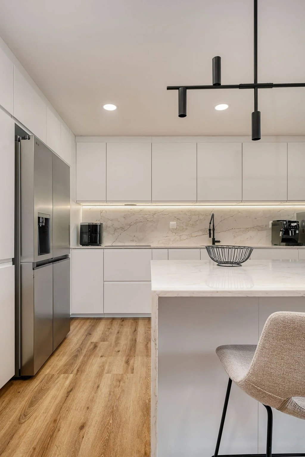 Cozinha moderna com armários brancos, parede revestida de mármore, eletrodomésticos de aço inox e bancada de mármore. Há uma pia preta e uma cadeira bege. Iluminação embutida e luminária linear preta no teto.