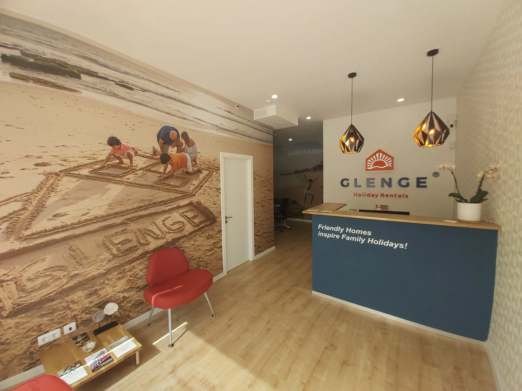 Projeto de Design de Interiores/Remodelação.
Balcão de recepção da Glenge Holiday Rentals com cor corporativa, logo em parede frontal, duas luminárias pendentes.
À esquerda, parede com impressão grande formato de uma família na praia, transmitindo o 