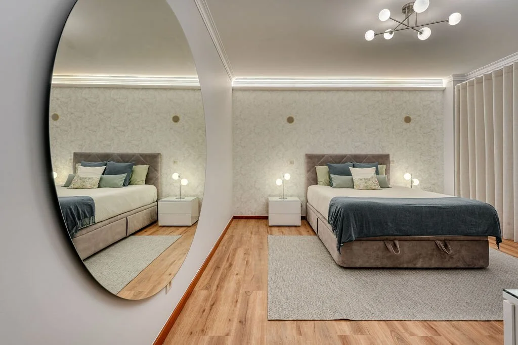 Quarto de casal visto através de um espelho na parede, com duas camas de solteiro, cada uma com uma cabeceira acolchoada, lençóis brancos, mantas e vários travesseiros em cores suaves. Há luminárias de mesa ao lado de cada cama, uma parede decorada c