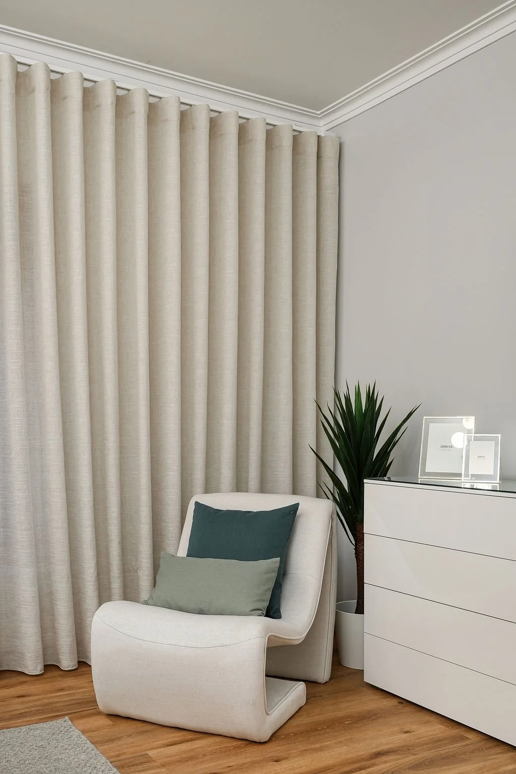 uma sala de estar com cortinas claras, uma poltrona branca com dois travesseiros (um verde escuro e outro cinza claro), uma planta alta ao lado de uma cômoda branca com molduras de fotos e duas luminárias de mesa.