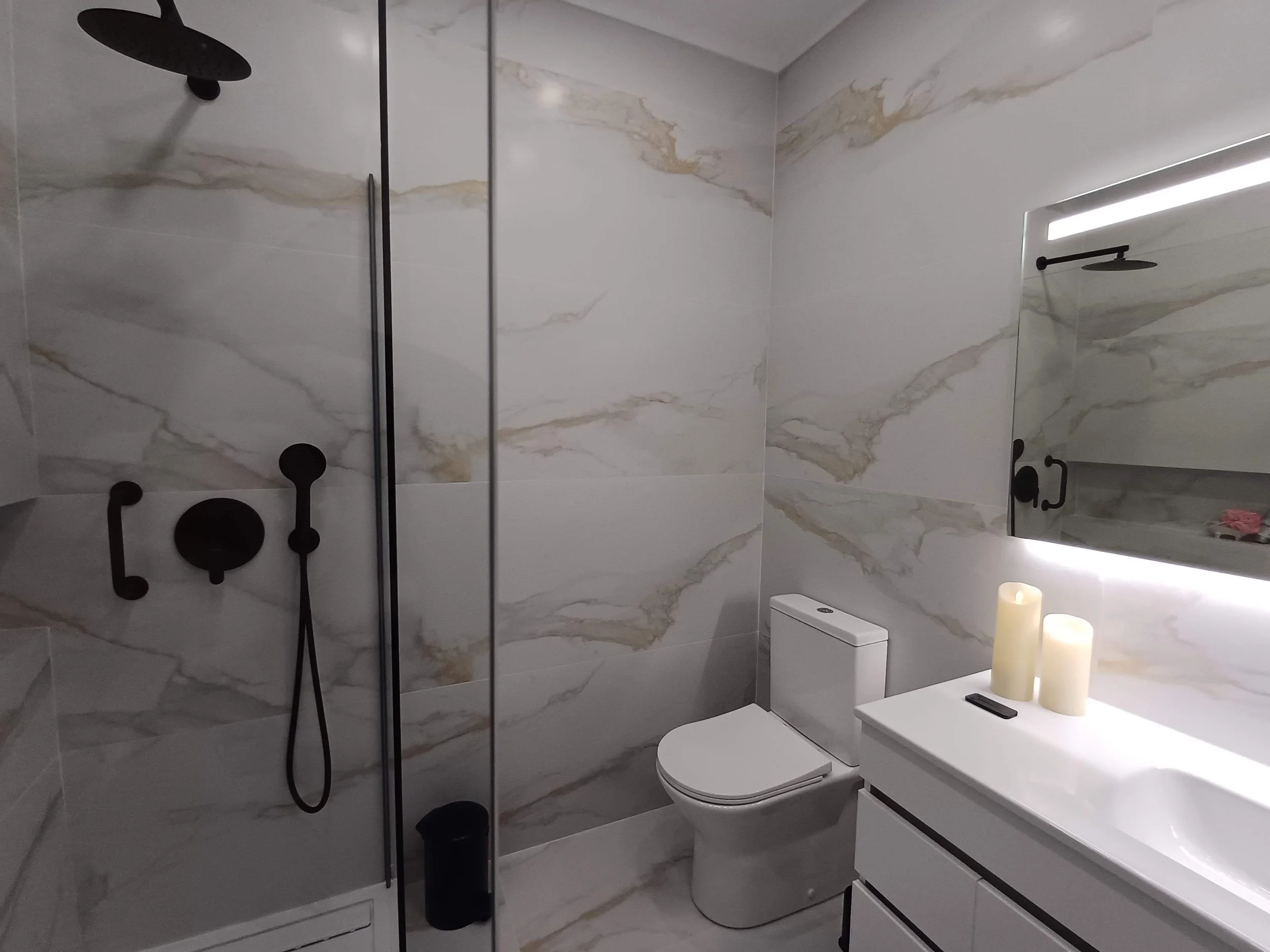 Banheiro moderno com paredes de mármore, ducha preta com acessórios, vaso sanitário branco e pia com espelho iluminado e velas