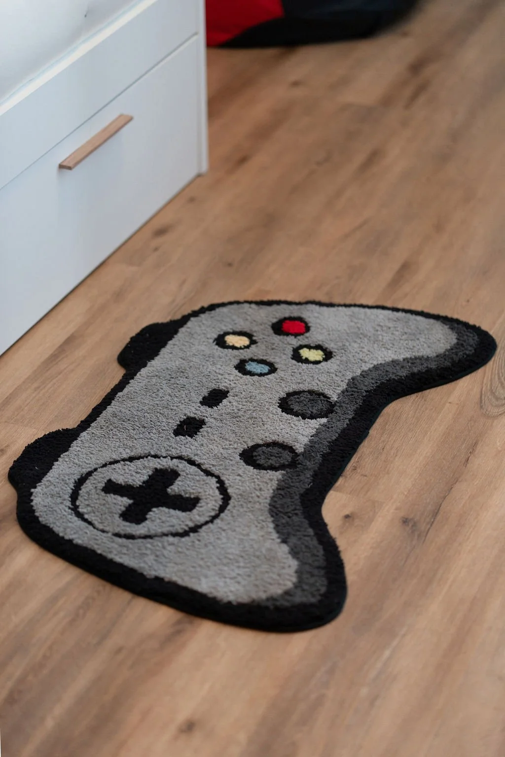 Tapete decorativo em forma de comando de vídeo-jogos, com detalhes coloridos, colocado no chão de madeira à saída de cama.