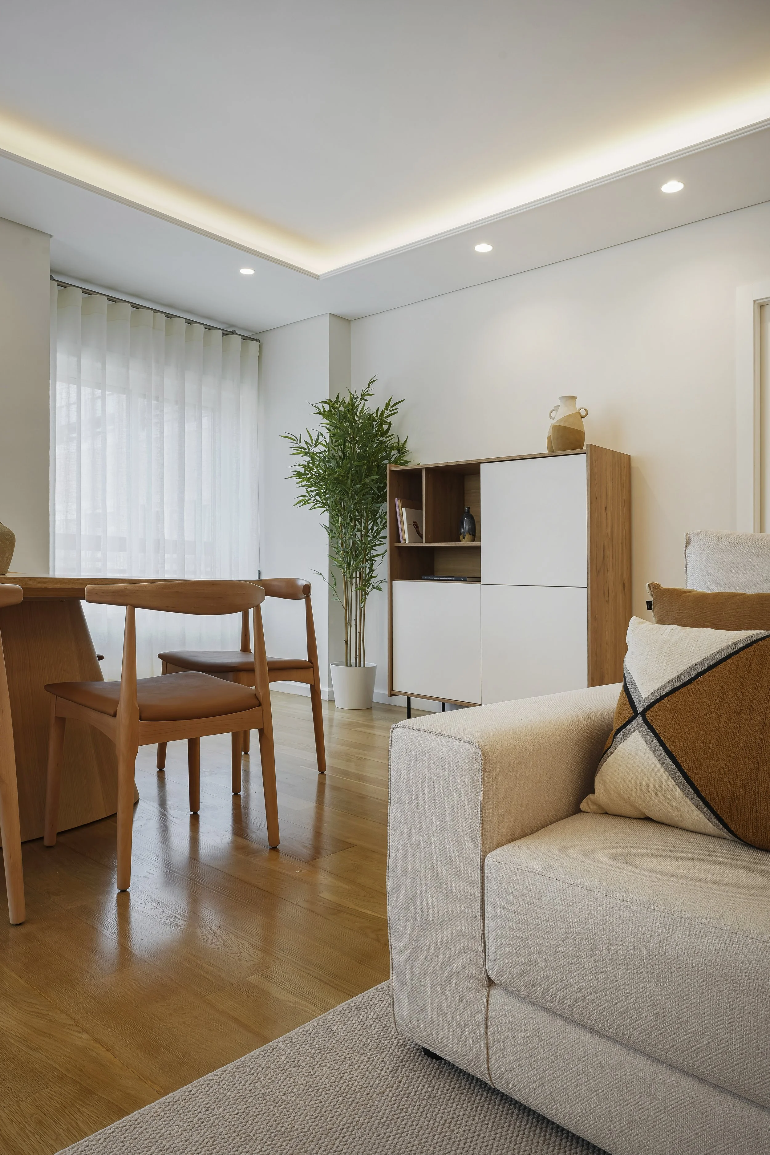 Sala de estar com móveis de madeira, sofá claro com almofada decorativa, prateleira de madeira com portas brancas, planta em vaso branco e janela com cortina de tecido branco.