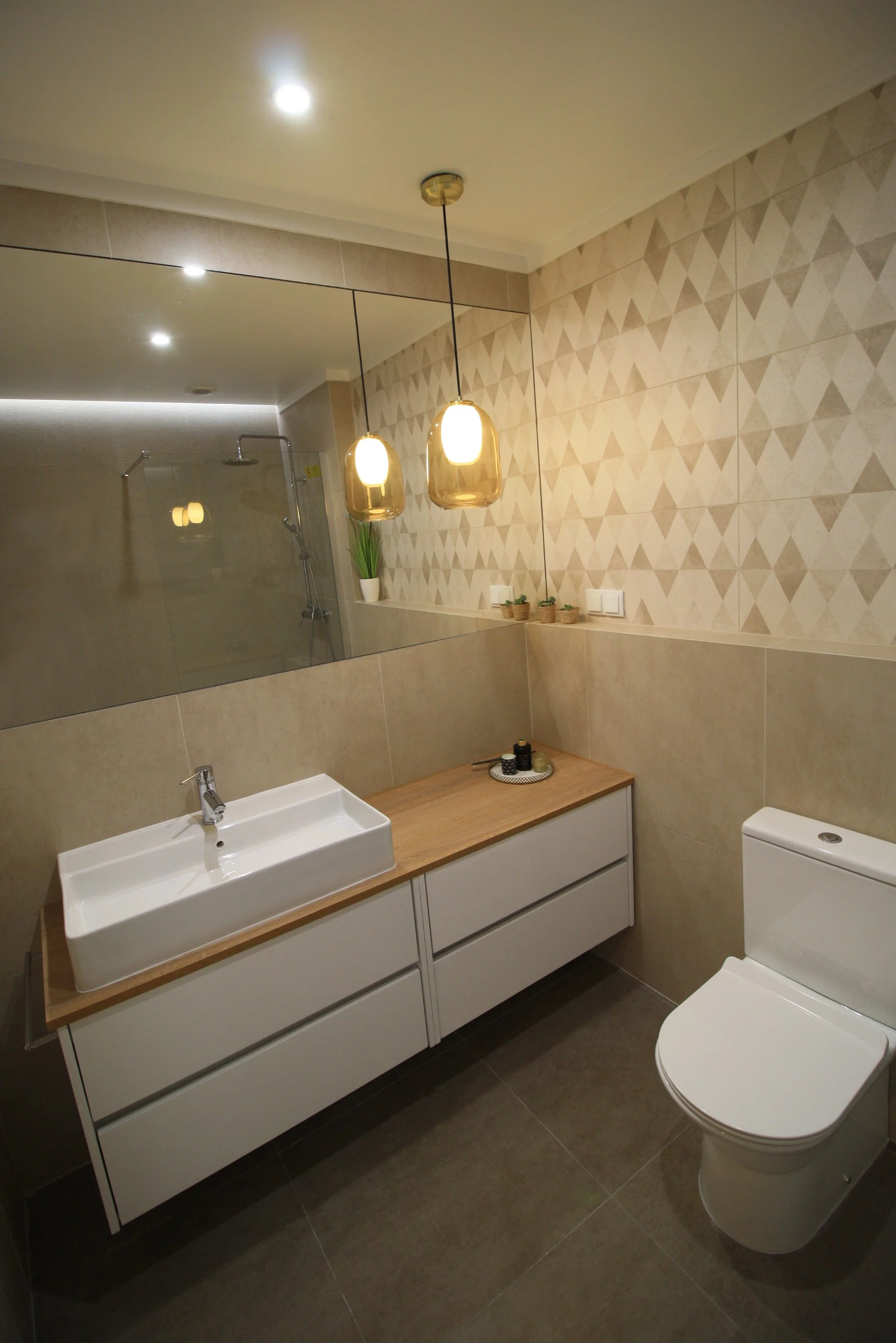 Banheiro moderno com pia de bancada de madeira, espelho grande, toalete branco, iluminação suspensa de lâmpadas douradas, parede decorada com azulejos geométricos, e se recebe uma ducha no box. Há também uma planta e algumas velas em um canto do banh