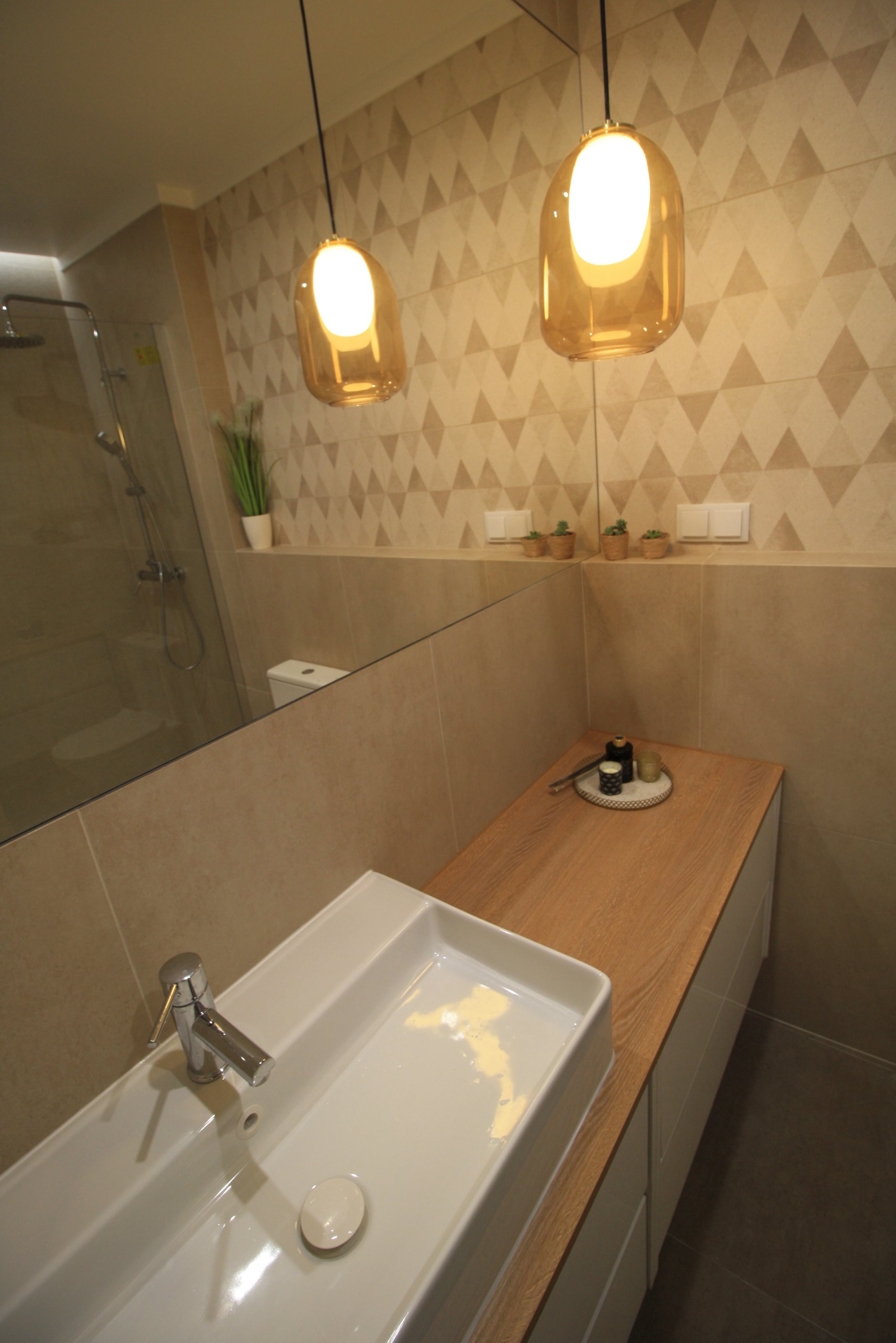 Banheiro com pia branca, espelho grande, luzes pendentes douradas, parede com azulejos de padrão triangular, bancada de madeira, decoração com plantas em mini vasos e objetos de banho pequenos.