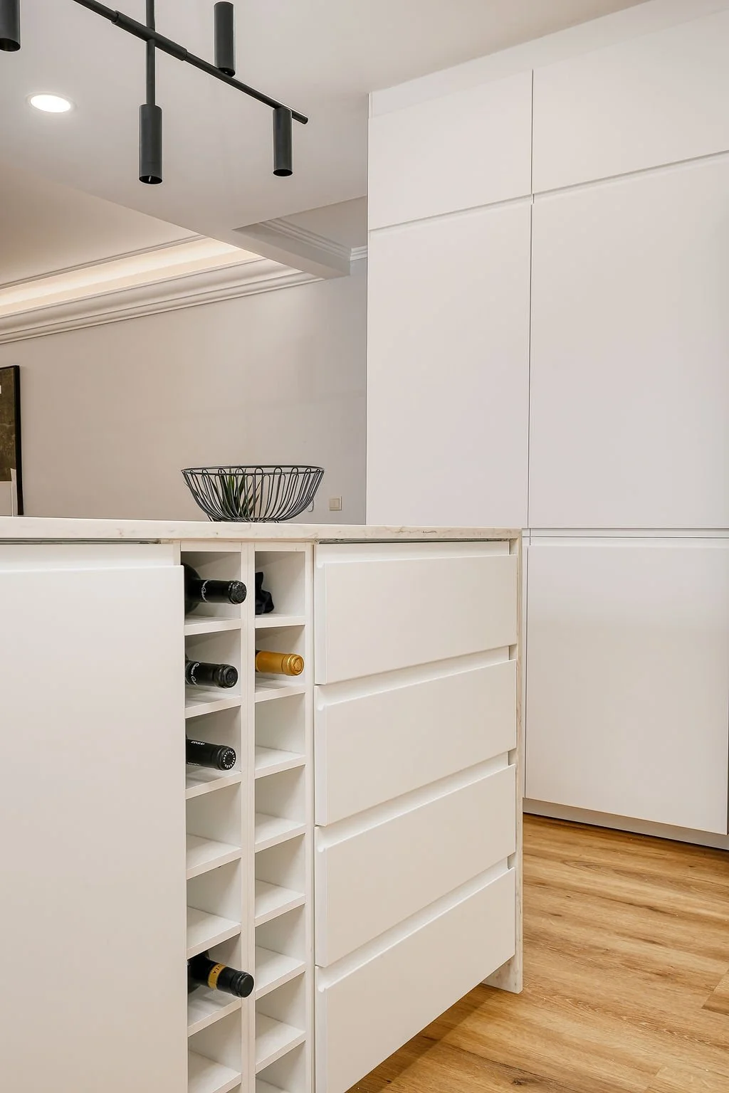 Cozinha moderna com armários brancos, bancada de mármore e um suporte para garrafas de vinho integrado na mobília, em piso de madeira.