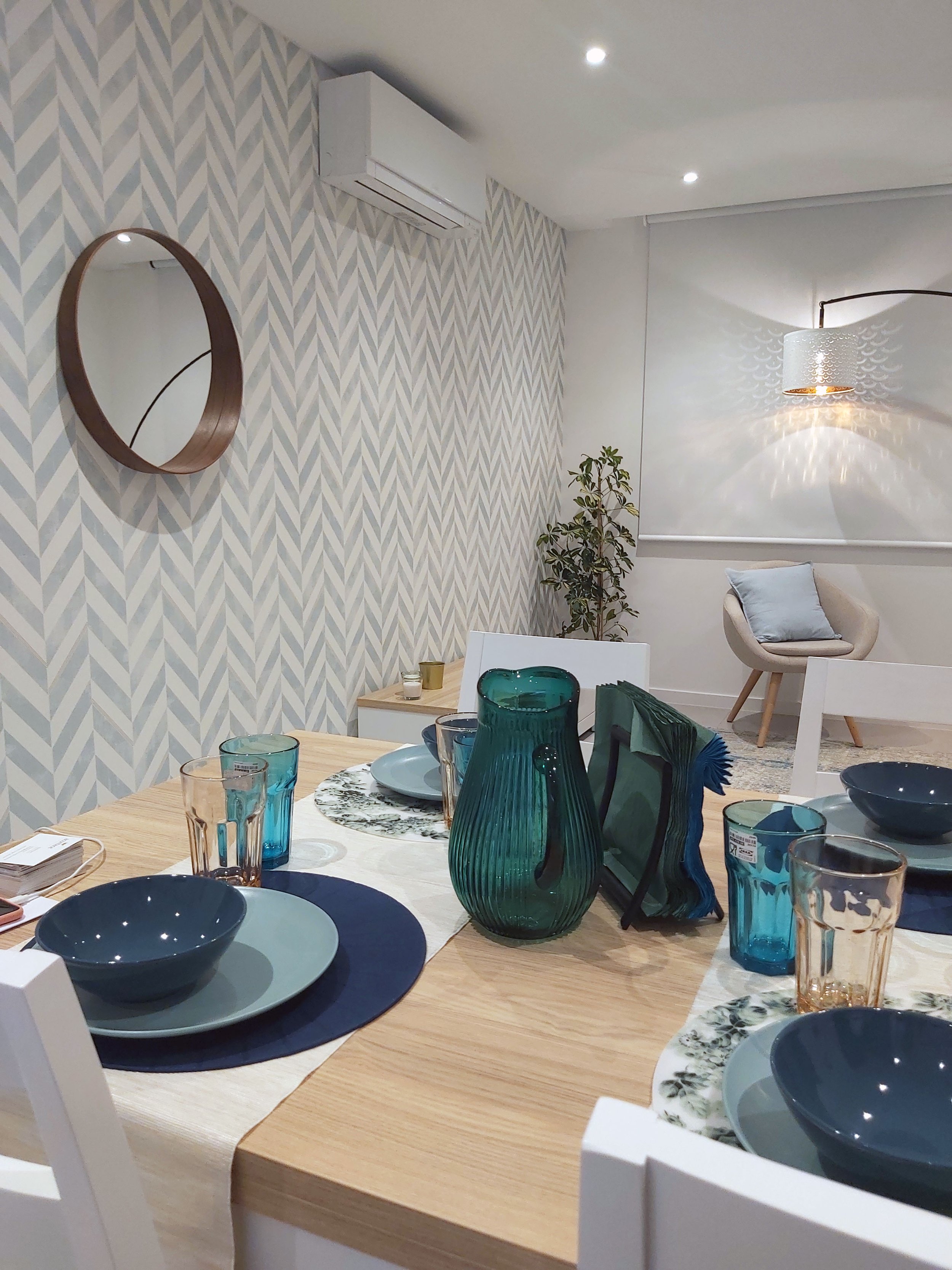 Reunião de jantar com mesa de madeira, pratos azuis, copos coloridos e várias garrafas de vidro verde. Sala decorada com espelho, lâmpada de parede, árvore de decoração e papel de parede com padrão geométrico.