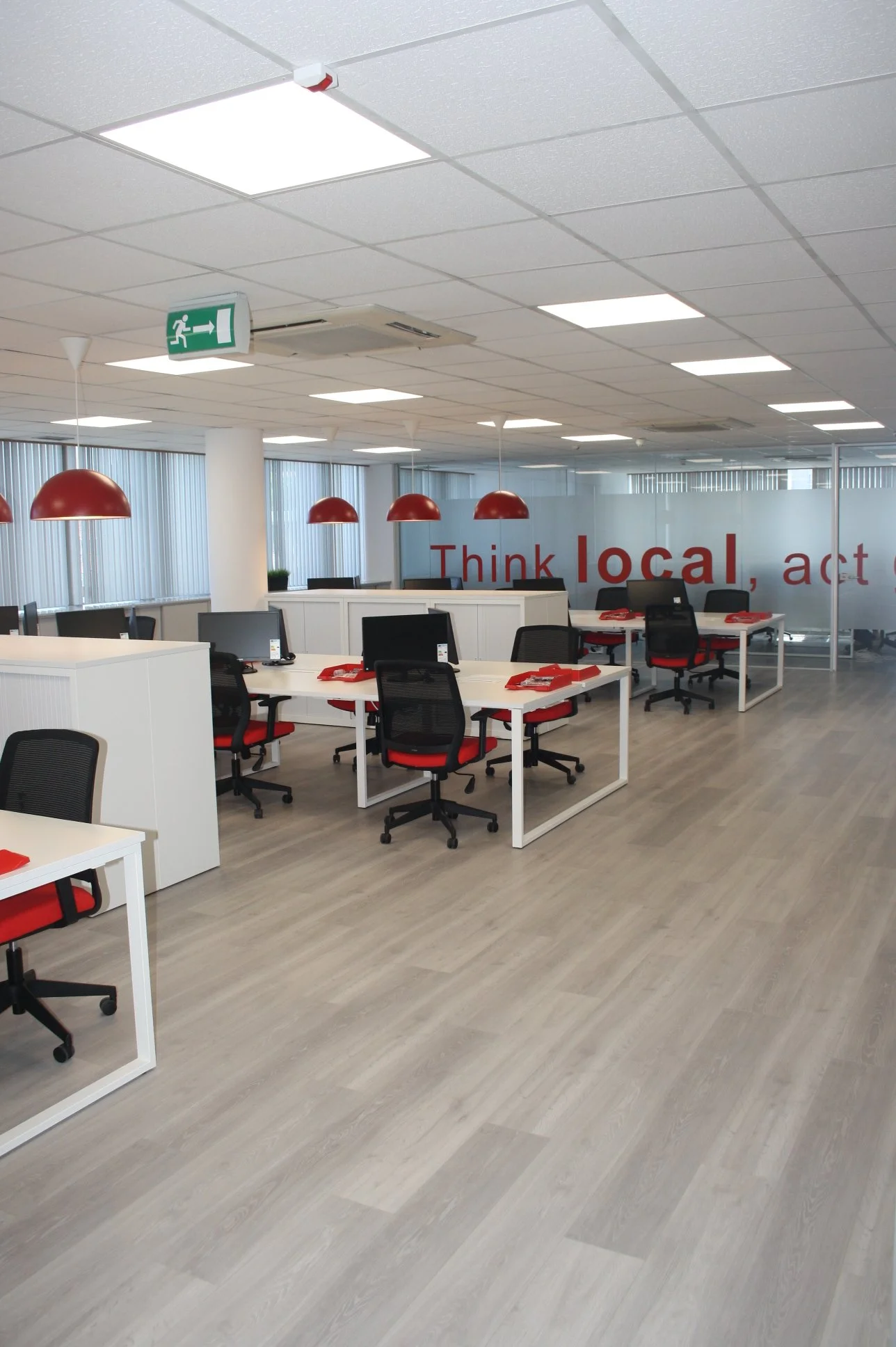 Escritório moderno com várias mesas de trabalho, cadeiras pretas com assentos vermelhos, luminárias vermelhas suspensas, piso de madeira clara, paredes de vidro e mensagem de incentivo na parede de vidro.