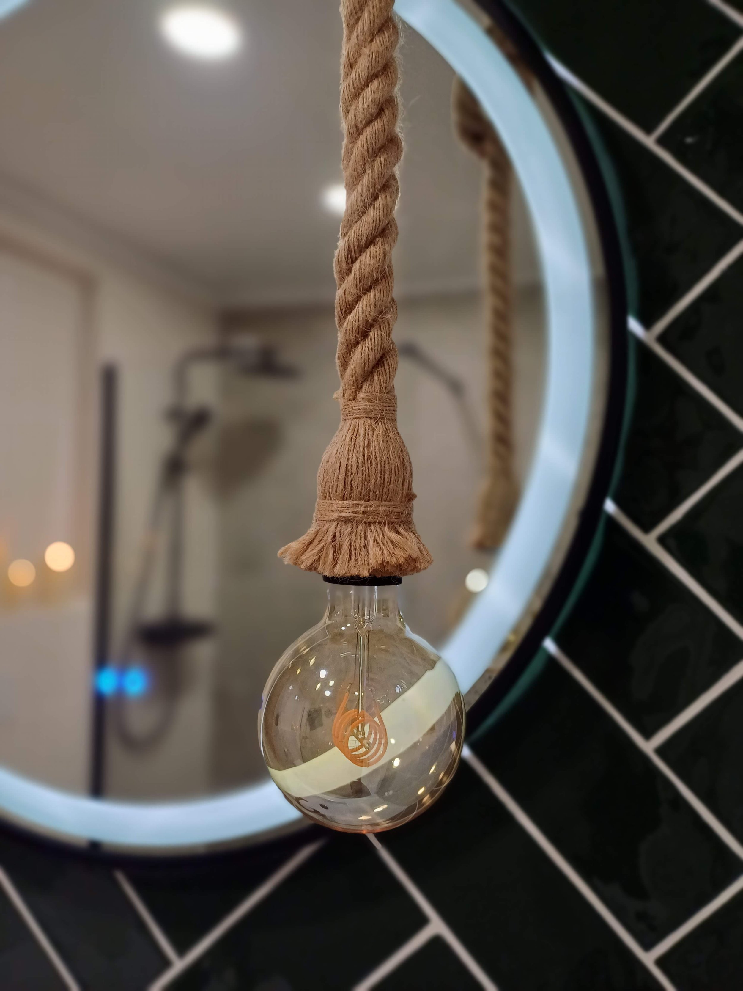 Lâmpada de filamento com pendurador de corda de sisal decorativa, refletida em um espelho redondo com moldura branca, no banheiro com azulejos pretos e brancos.