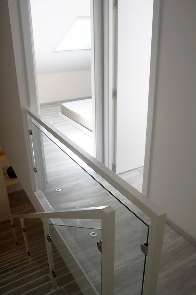 Corredor com sacada de vidro, visualizando um quarto com cama e janela de teto.