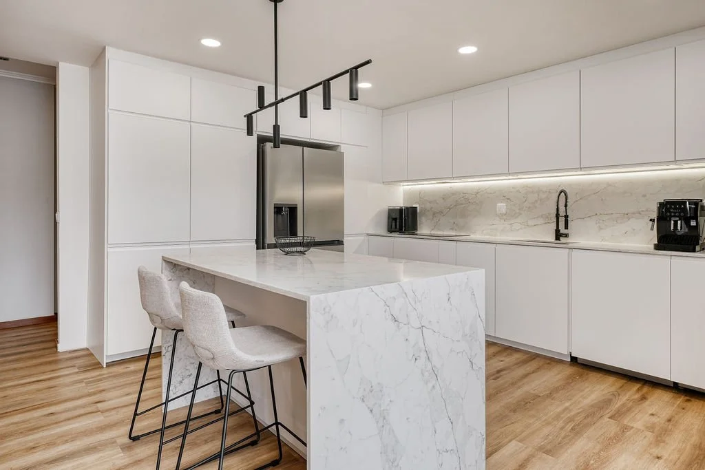 Cozinha moderna com armários brancos, ilha de mármore branco, eletrodomésticos de aço inox, iluminação embutida e piso de madeira laminada.