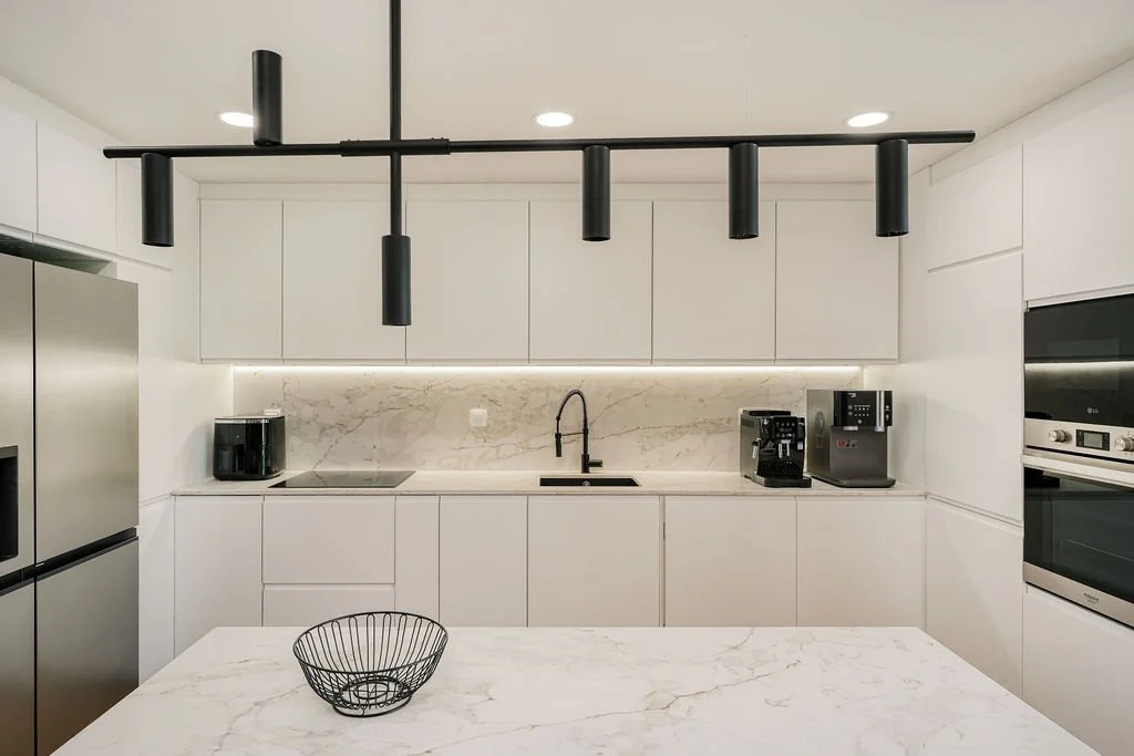 Cozinha moderna com armários brancos, bancada de mármore, eletrodomésticos em aço inox, balcão com bule de café, e uma mesa com uma tigela de arame