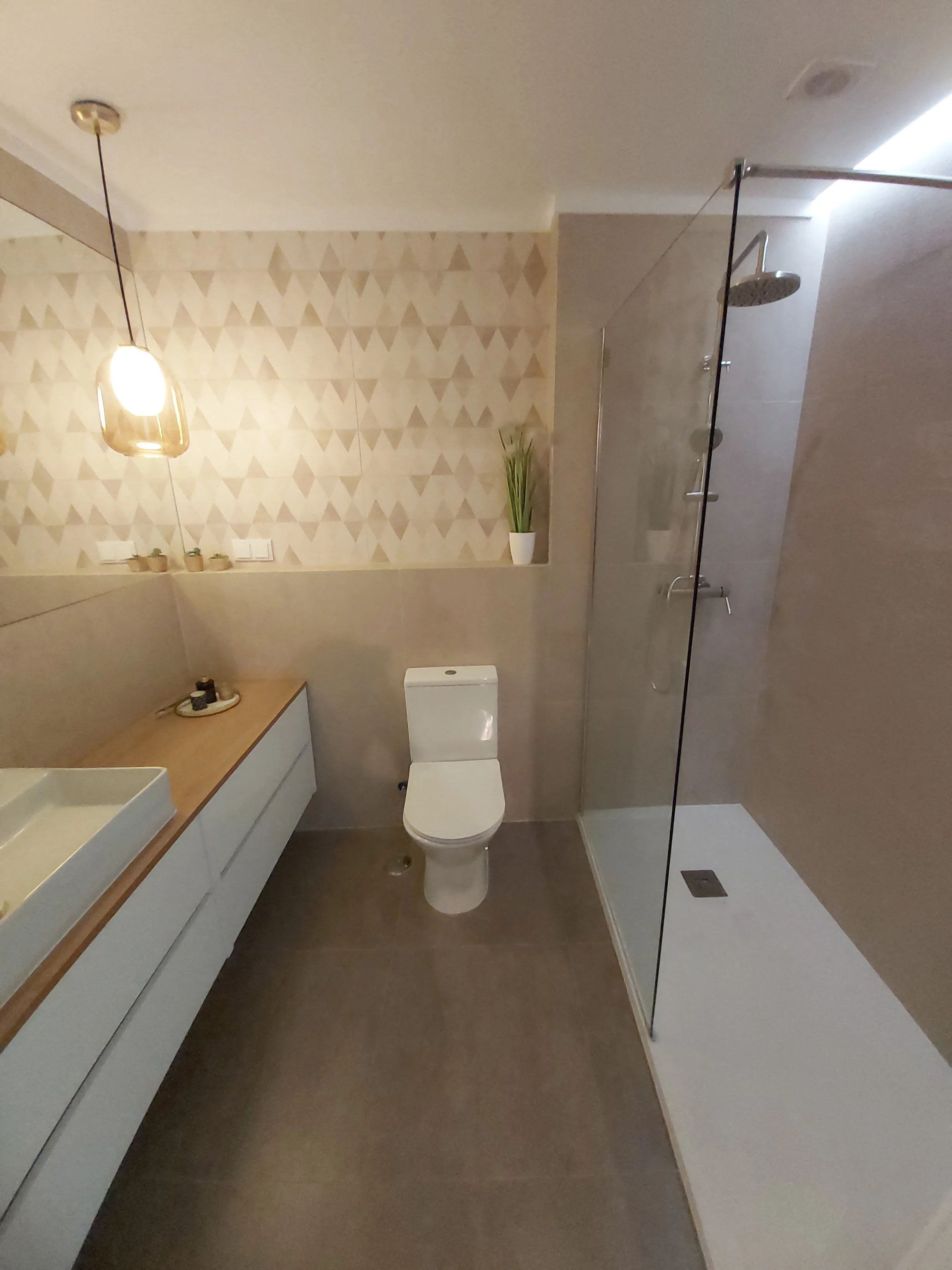Banheiro com vaso sanitário, pia com balcão de madeira, parede com revestimento de azulejos decorativos, ducha de vidro, luz pendente e uma planta no canto.