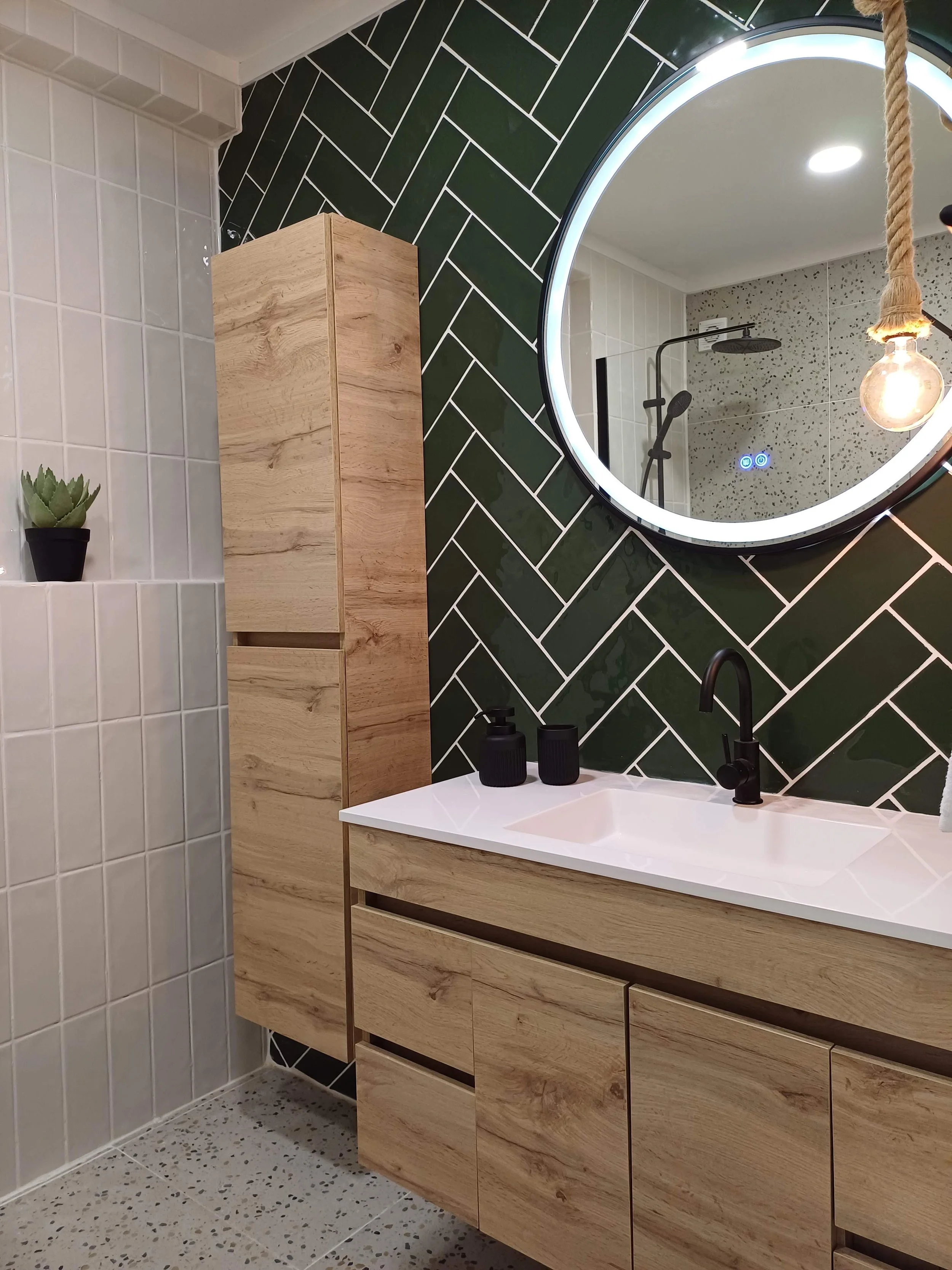 Banheiro com espelho redondo com iluminação LED, lavatório de bancada de madeira, paredes de azulejo, torneira preta, acessórios pretos, planta em vaso, parede de azulejo verde escuro em padrão diagonal.