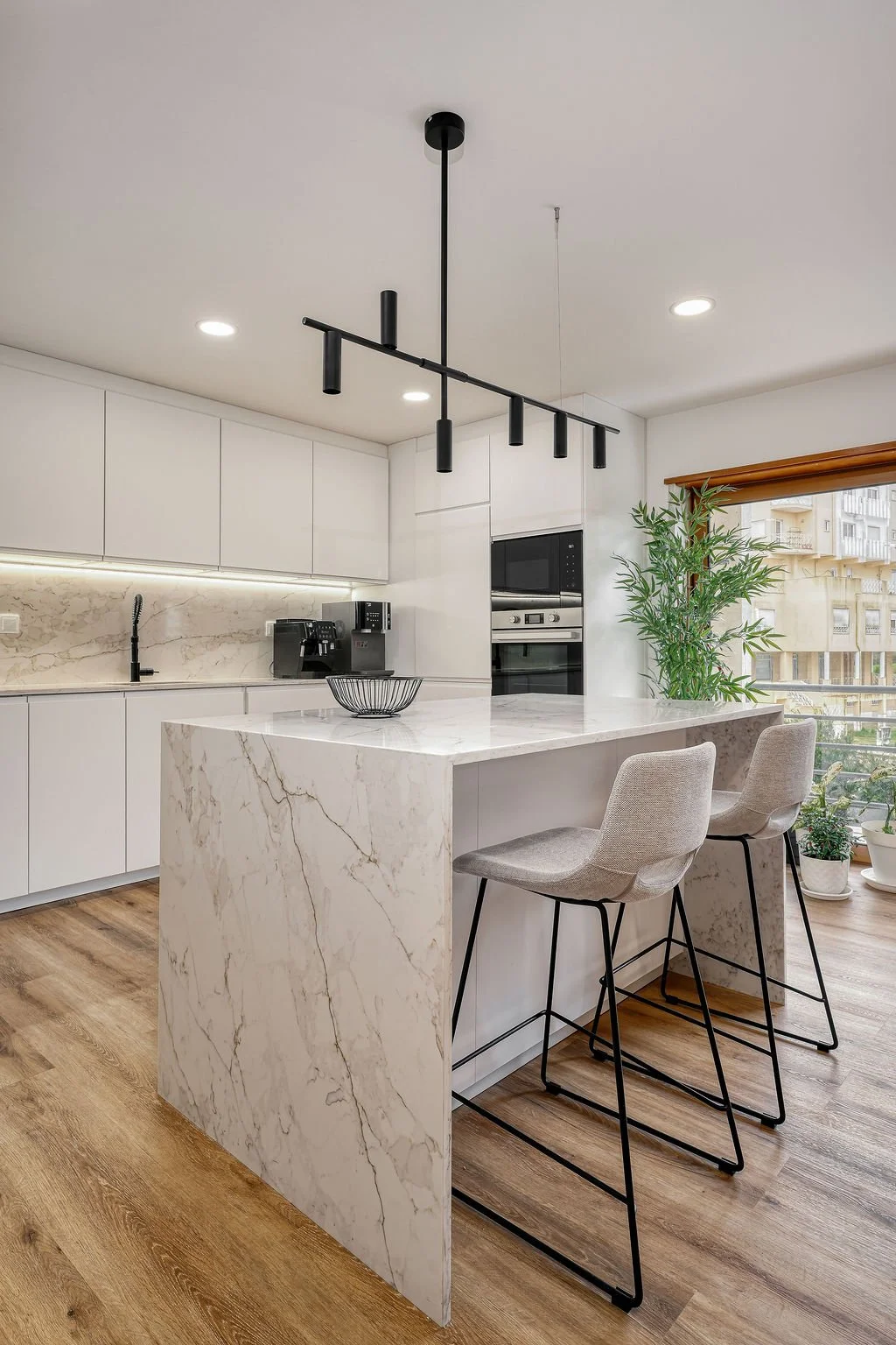 Cozinha moderna com ilha de mármore branco, duas cadeiras altas de almofada, armários brancos, eletrodomésticos embutidos, plantas na varanda e iluminação suspensa preta.