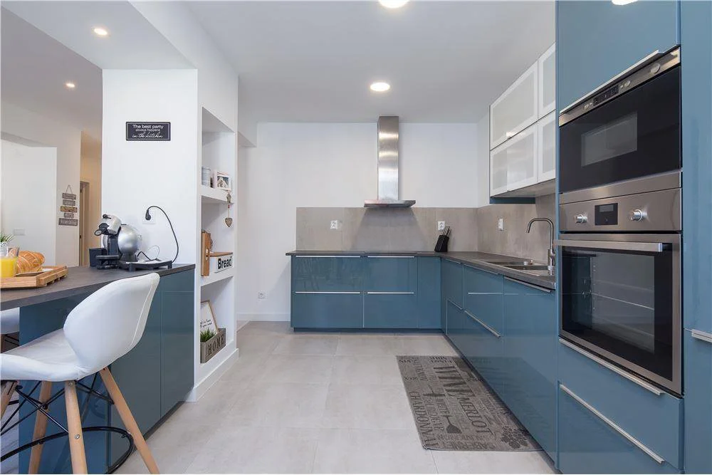 Projeto de Design de Interiores e Remodelação de Apartamento em Carnaxide, Oeiras.
Perspetiva de Cozinha contemporânea com armários azuis petróleo, bancada de granito, Península com quatro cadeiras altas, eletrodomésticos embutidos e iluminação de LE