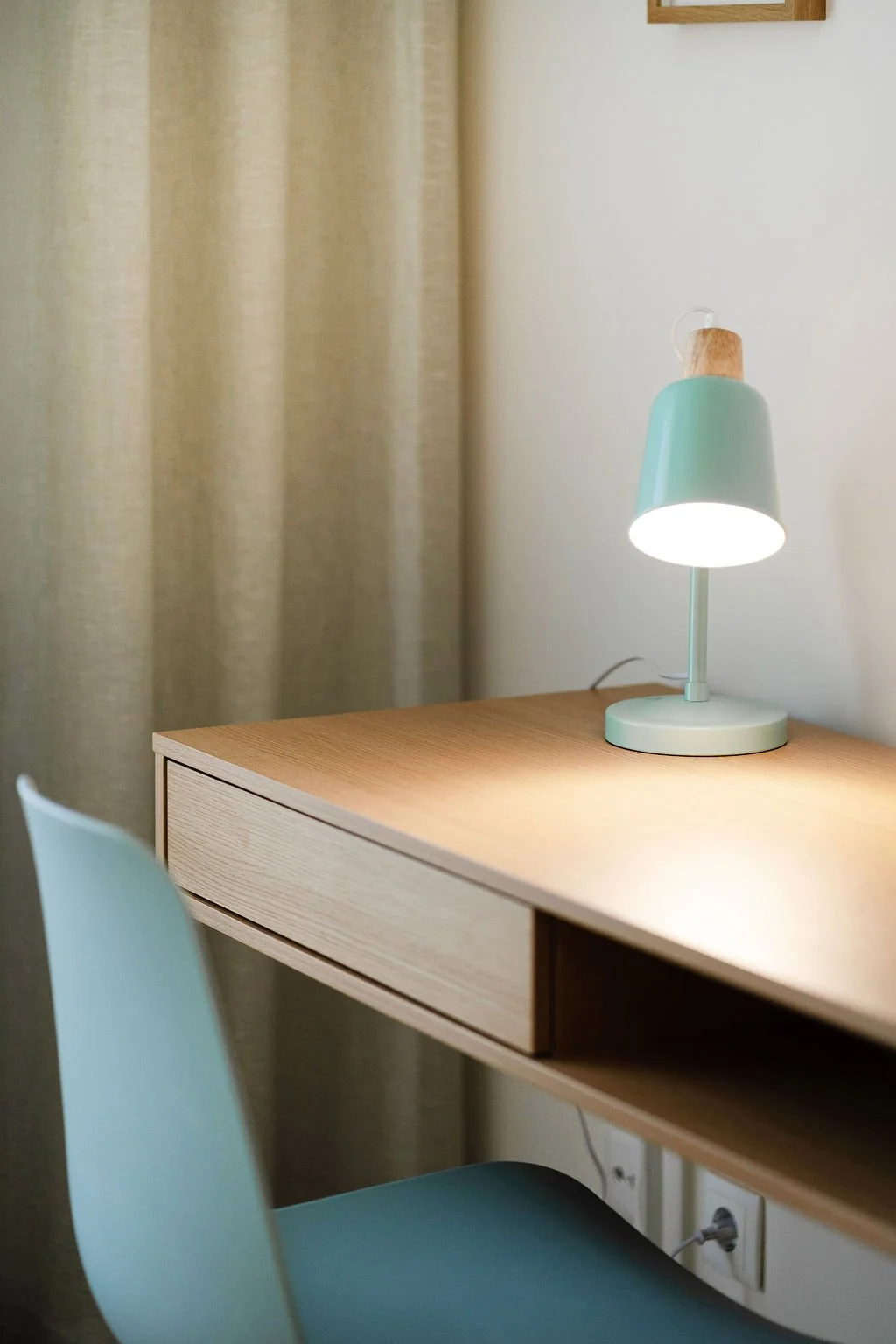Mesa de madeira clara com uma gaveta, uma cadeira azul na frente e uma luminária de mesa azul acesa ao lado de uma tomada elétrica.
