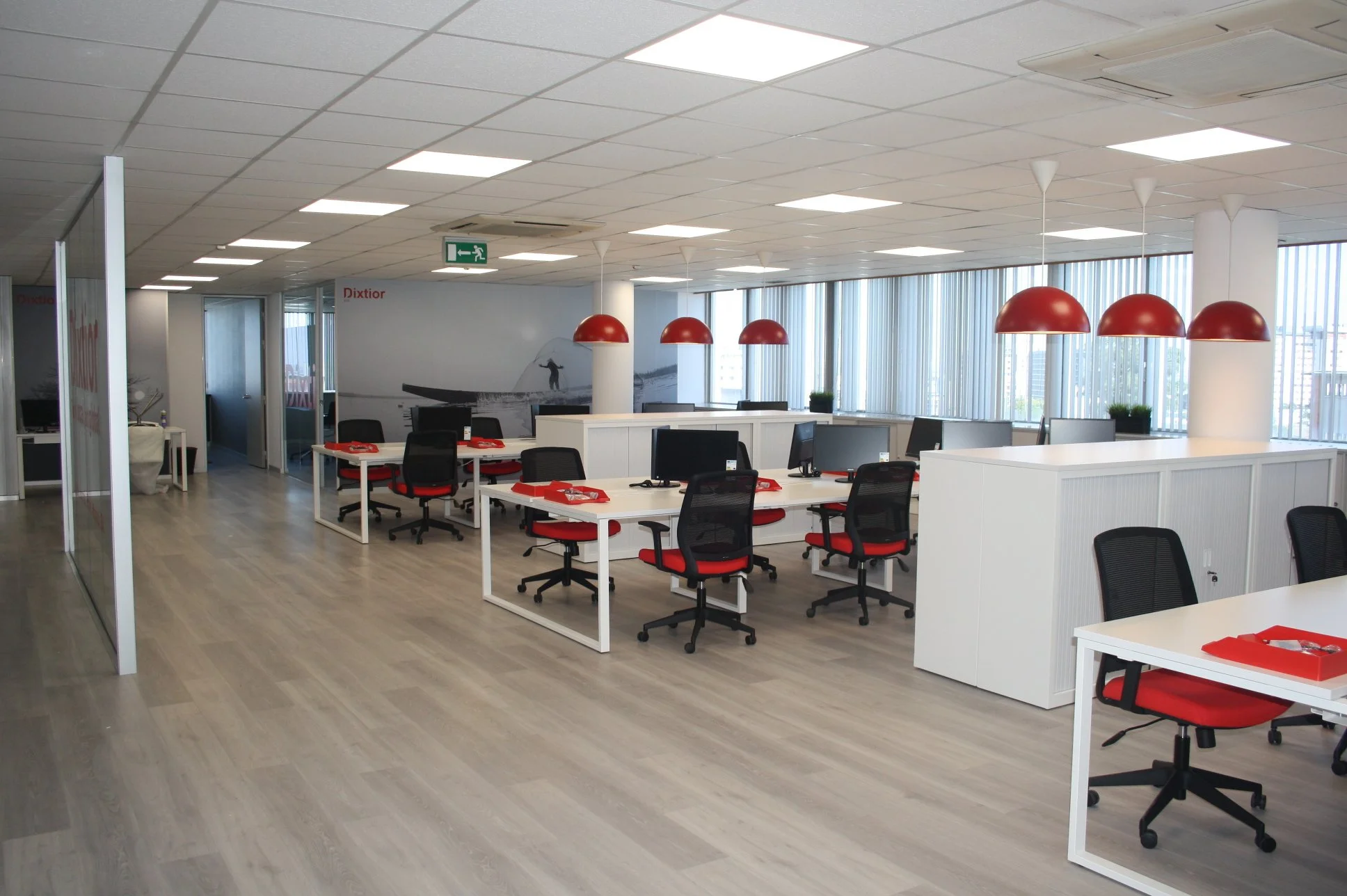 Escritório moderno com mesas de trabalho, cadeiras pretas e vermelhas, luminárias vermelhas pendentes, piso de madeira clara e janelas grandes com cortinas de vidro.