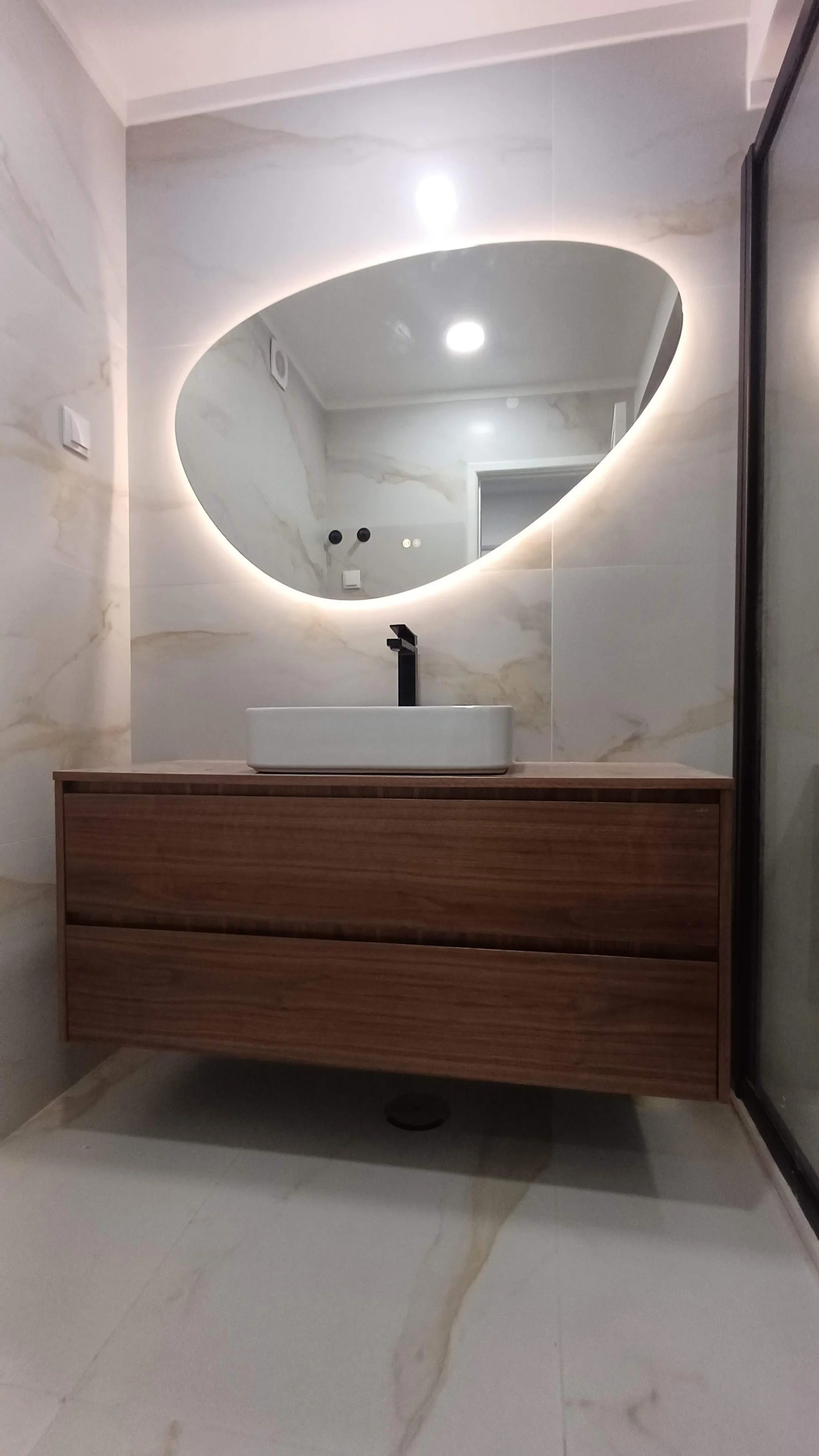 Espelho irregular com iluminação de LED ao redor, móvel de madeira com pia branca e torneira preta, parede de mármore claro, piso de mármore claro, porta de vidro ao lado.