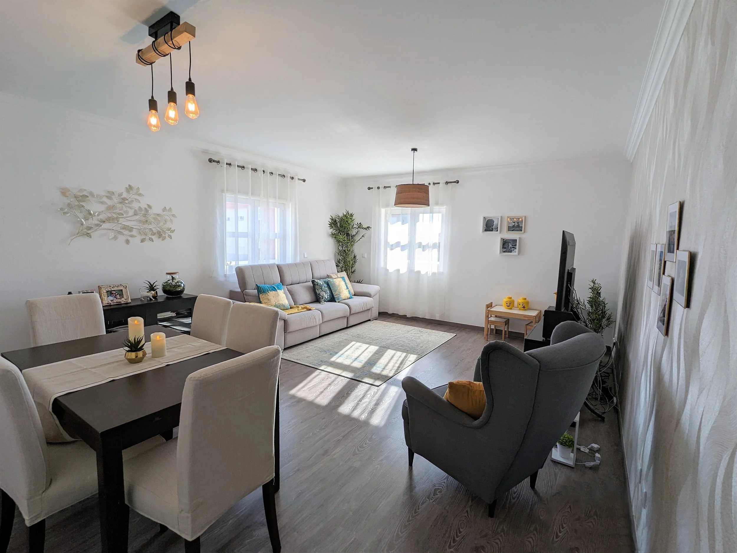 Sala de estar com sofá bege, poltrona cinza, mesa de jantar com seis cadeiras brancas, televisão com suporte, janelas com cortinas brancas, parede decorada com quadros e plantas.