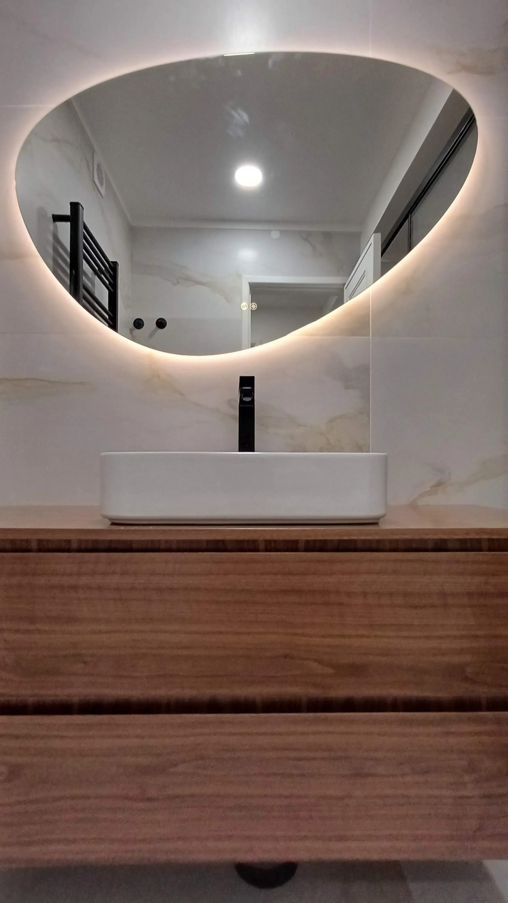 Espelho redondo com luz de LED ao redor acima de uma pia branca, instalada em uma bancada de madeira clara no banheiro.