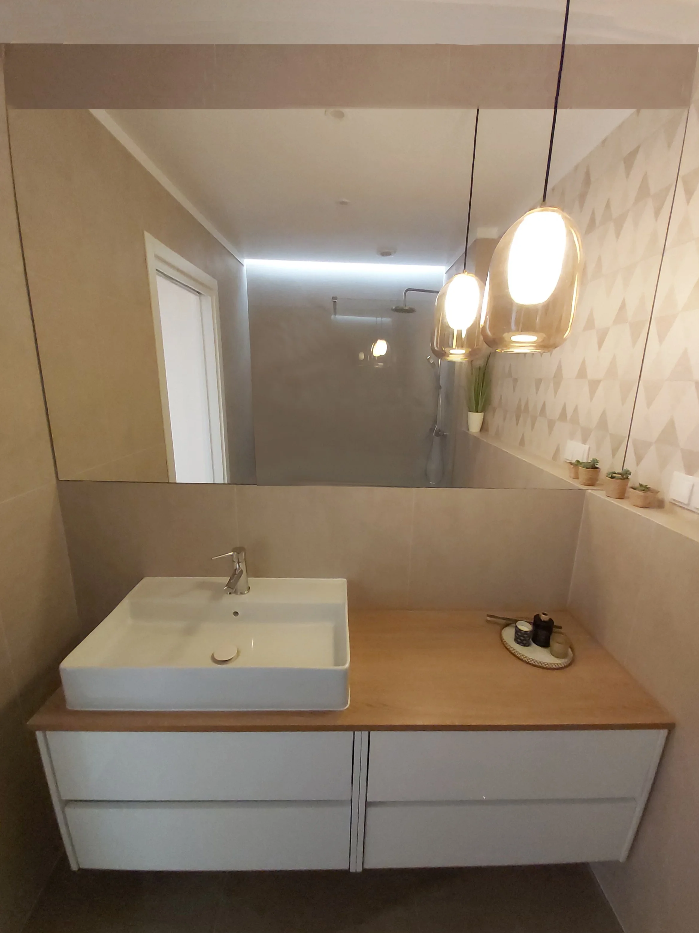 Espelho grande no banheiro refletindo a pia, luzes pendentes, parede decorativa, balcão de madeira com itens decorativos, vaso com planta ao fundo.