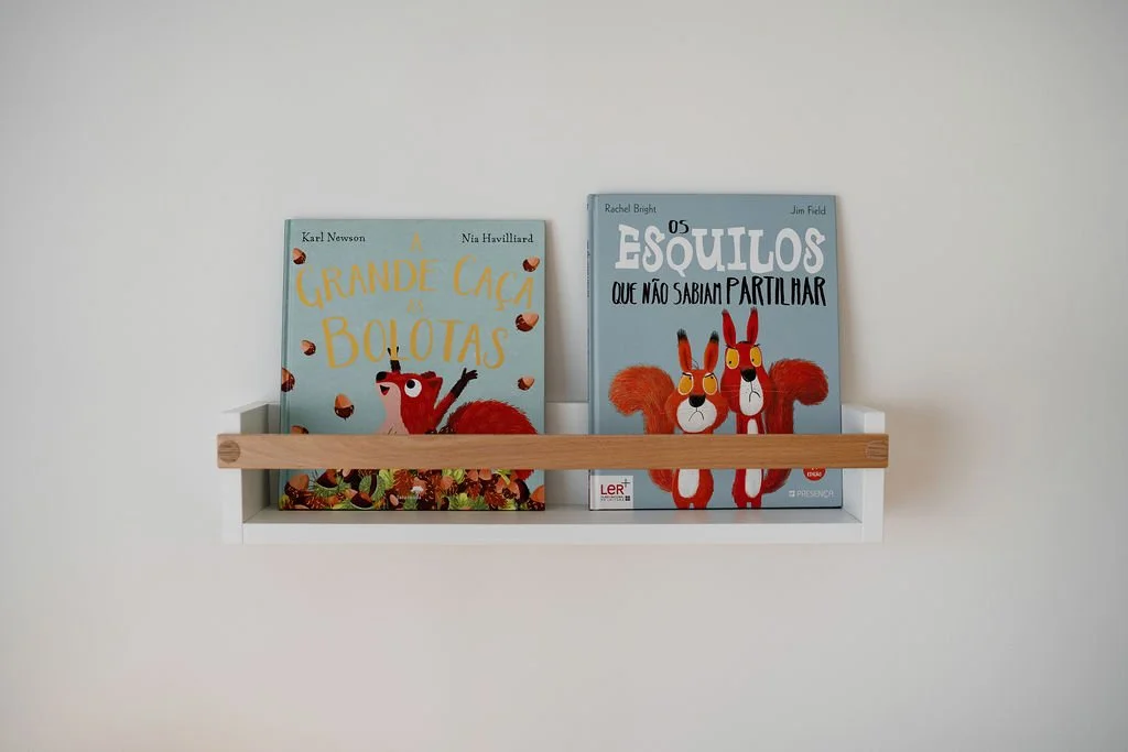 Prateleira branca com uma faixa de madeira na parede, segurando dois livros infantis com ilustrações de esquilos vermelhos.
