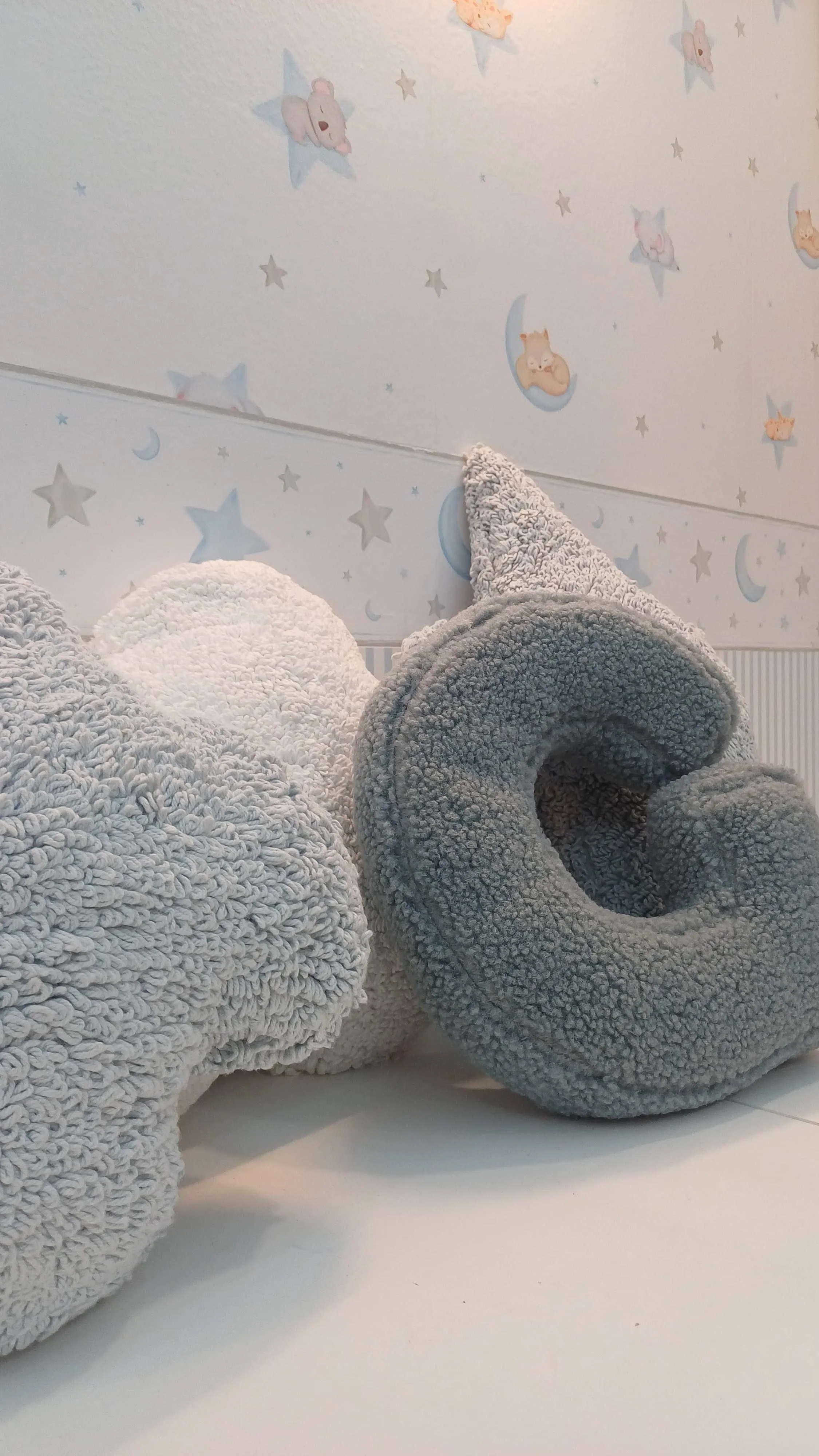 Três objetos de decoração de crochê em cores cinza e branco com fundo de parede decorada com desenhos de animais em estrelas e luas.