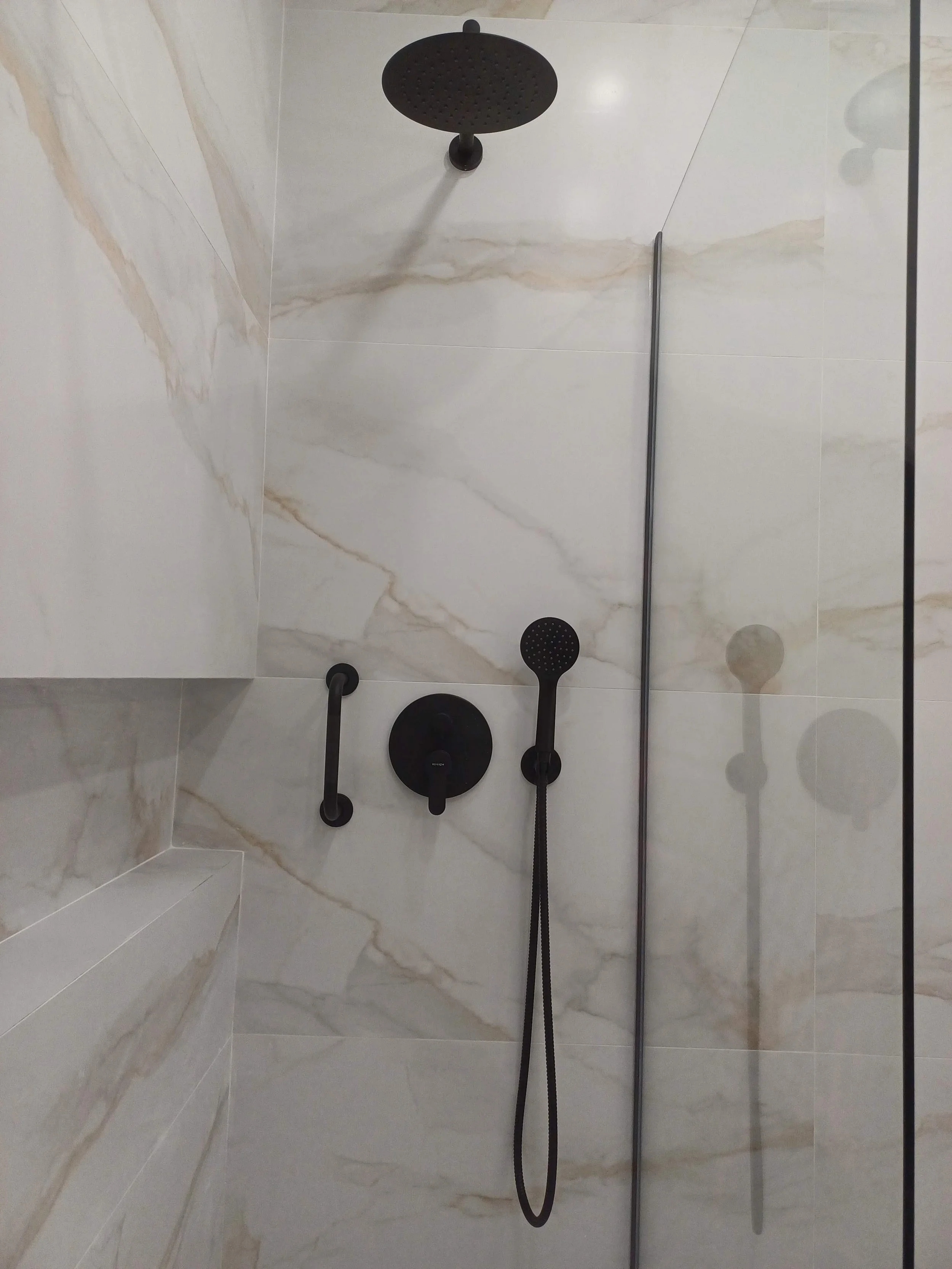 Chuveiro de banho com revestimento em mármore, painel de controle preto, ducha fixa redonda e ducha de mão, todos em cor preta, e uma divisória de vidro ao lado.