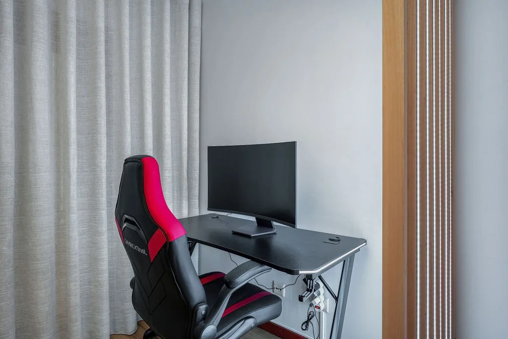 Escritório com cadeira gamer preta e rosa, mesa de computador com monitor e remo, parede clara, cortinas bege, parede de madeira com ripas.